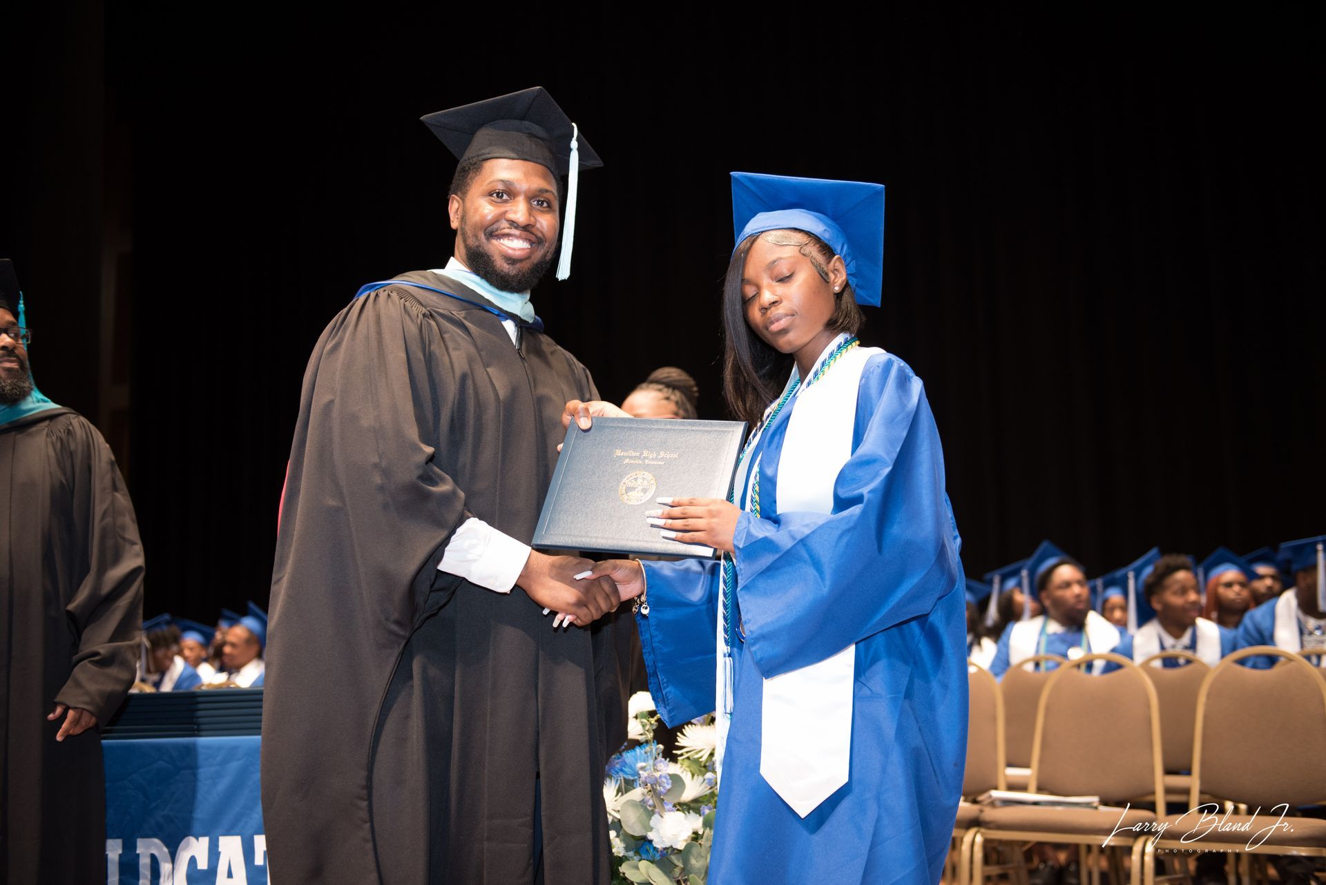 Person in black graduation attire hands diploma to person in blue graduation attire on stage.