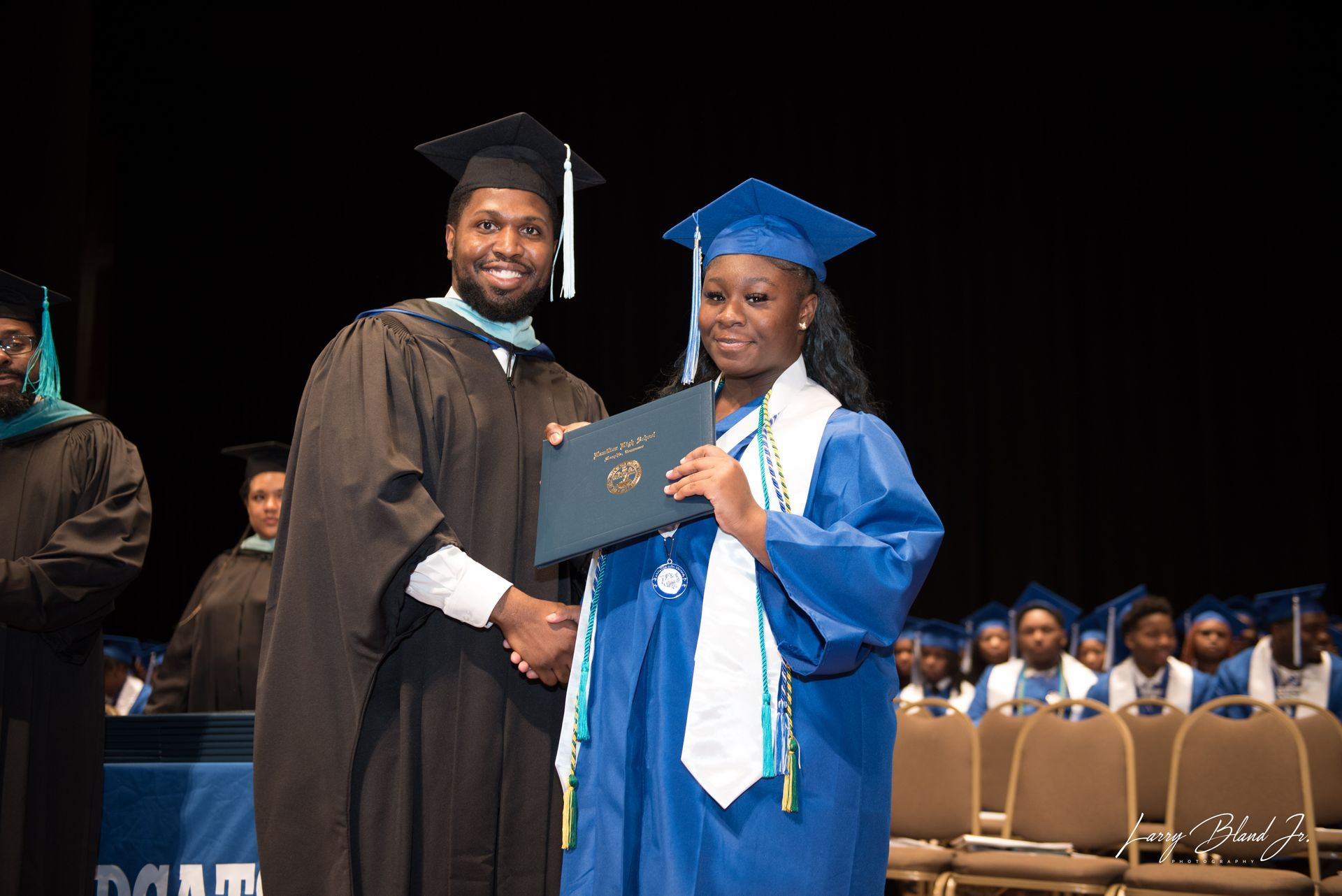 Man in black robe hands diploma to person in blue gown at graduation.