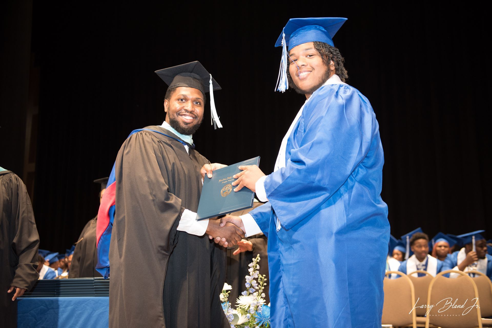 Man in black gown hands diploma to person in blue gown onstage.