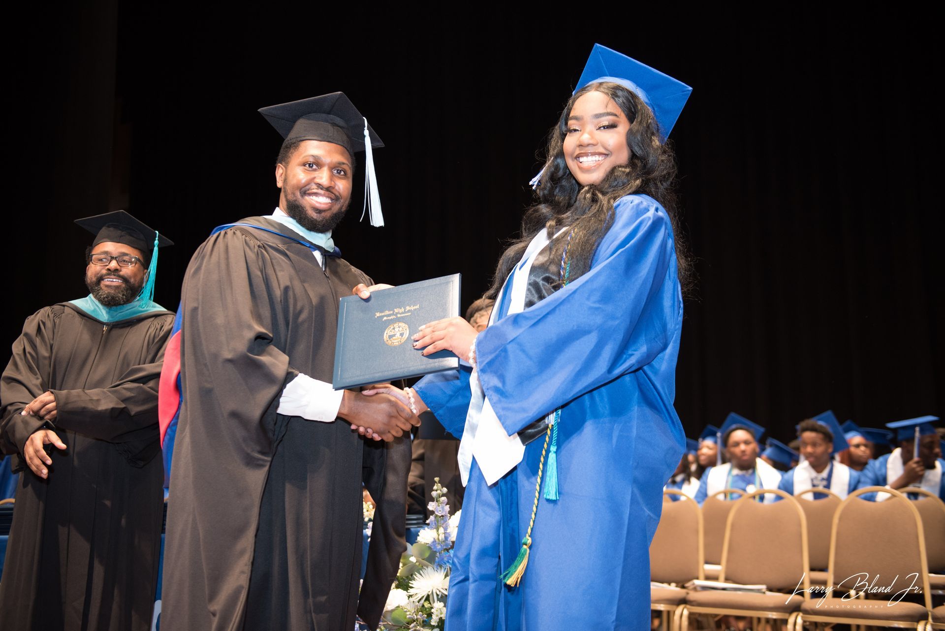 Person in black robe hands diploma to person in blue graduation gown on a stage.