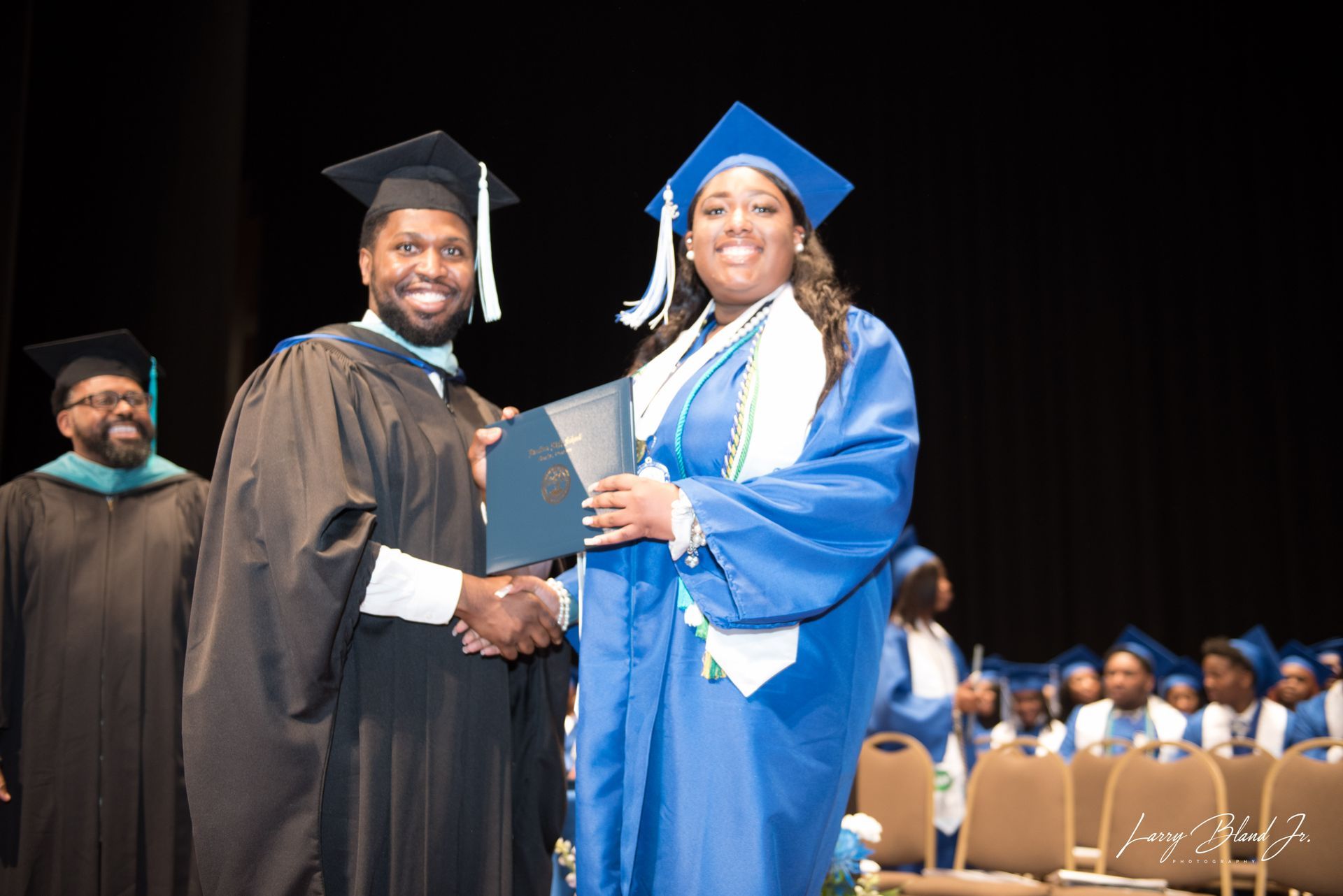 A graduate in blue robe shaking hands, receiving a diploma from a person in a black gown at a graduation ceremony.