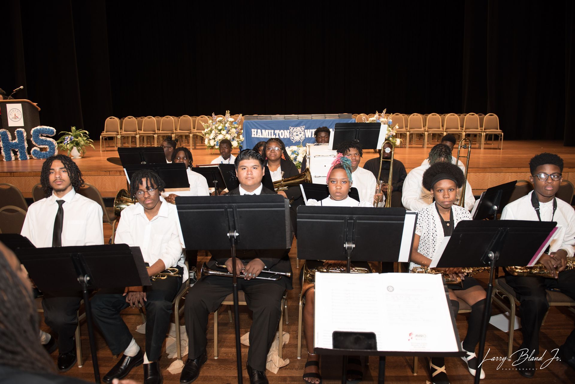 School band performing on stage, wearing formal attire, sheet music displayed on stands.