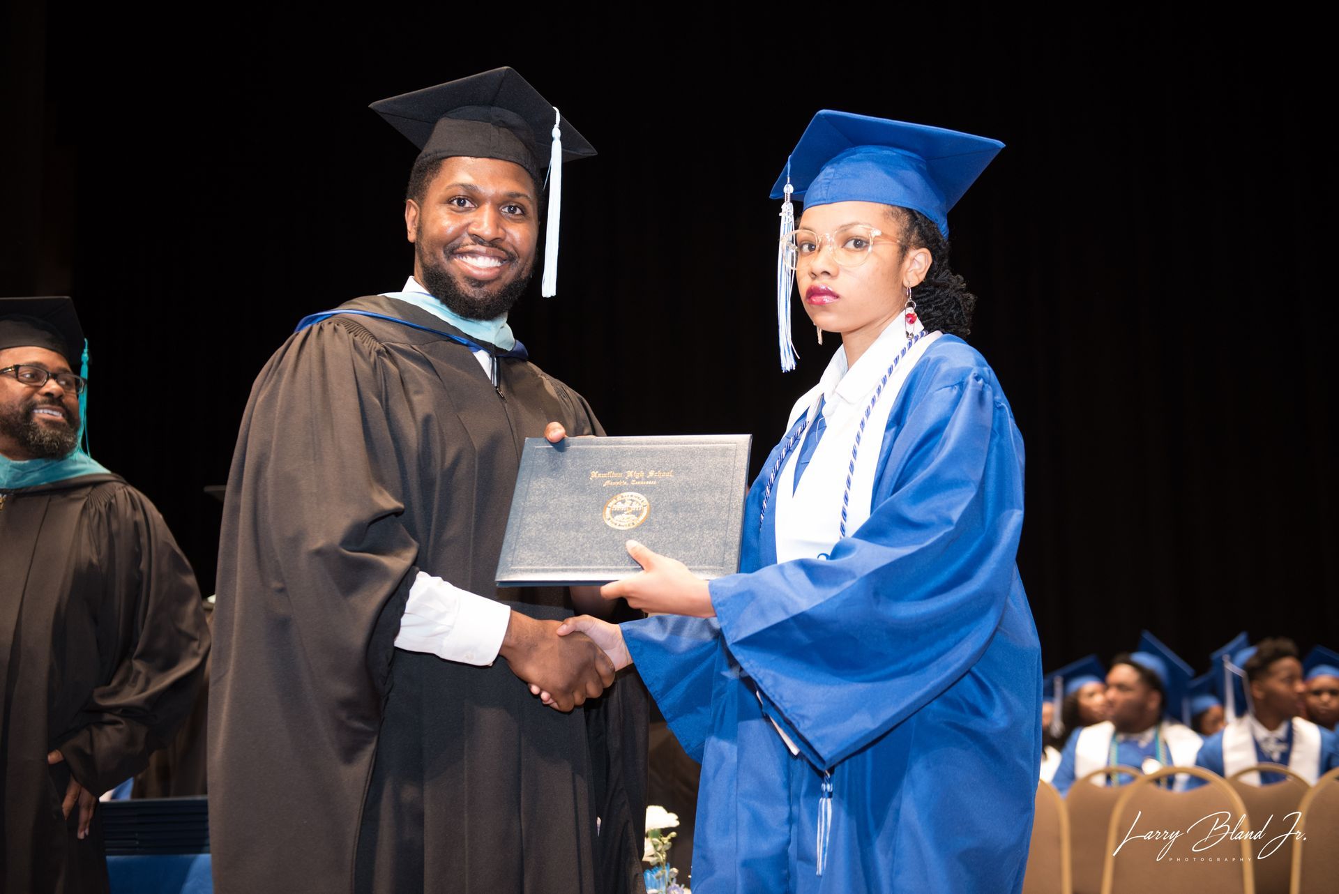Man in black gown hands a diploma to a person in a blue gown at a graduation ceremony.
