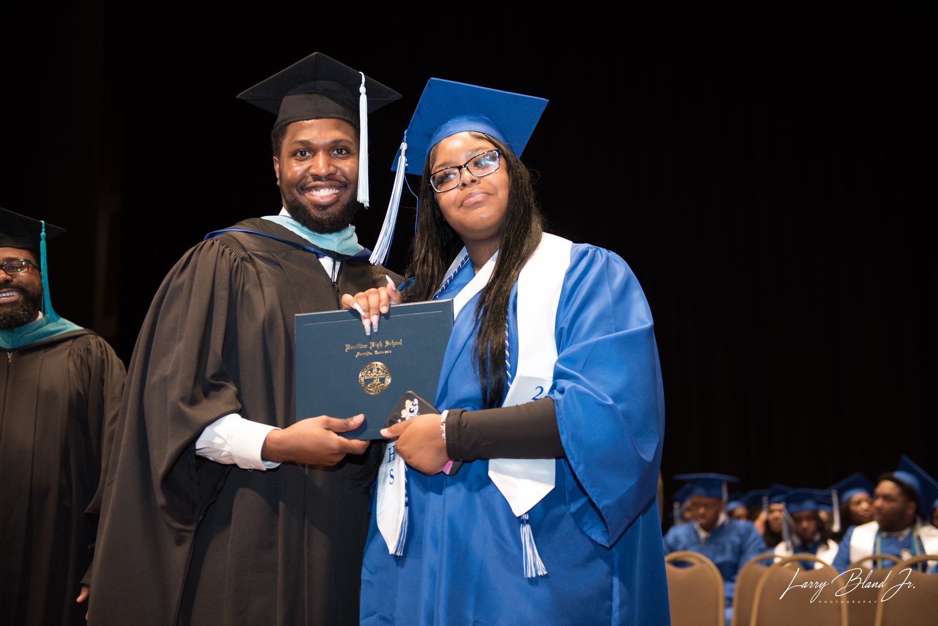 Person in black graduation gown hands diploma to person in blue gown at ceremony.