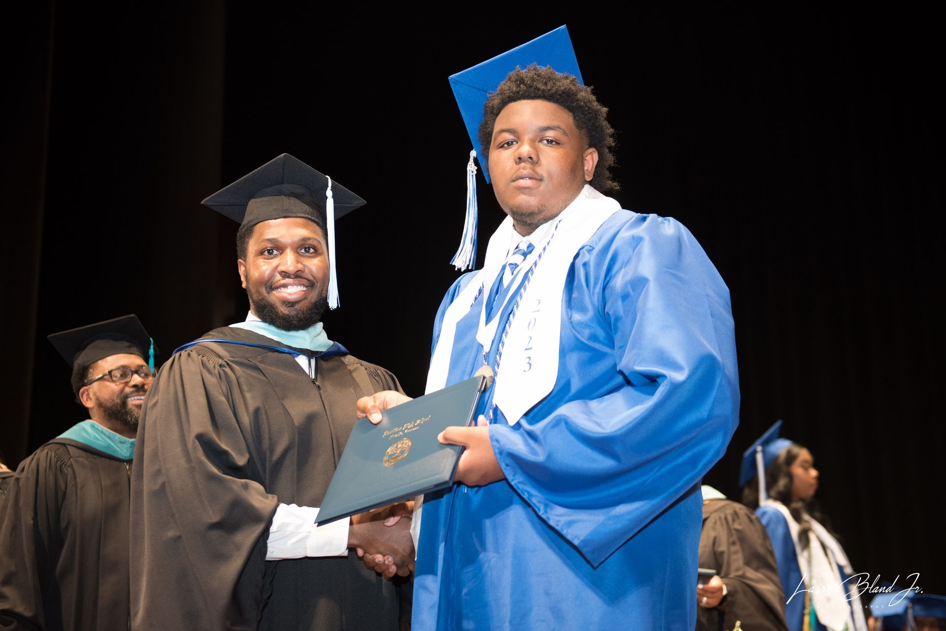 Man in graduation gown hands diploma to student in blue gown onstage.