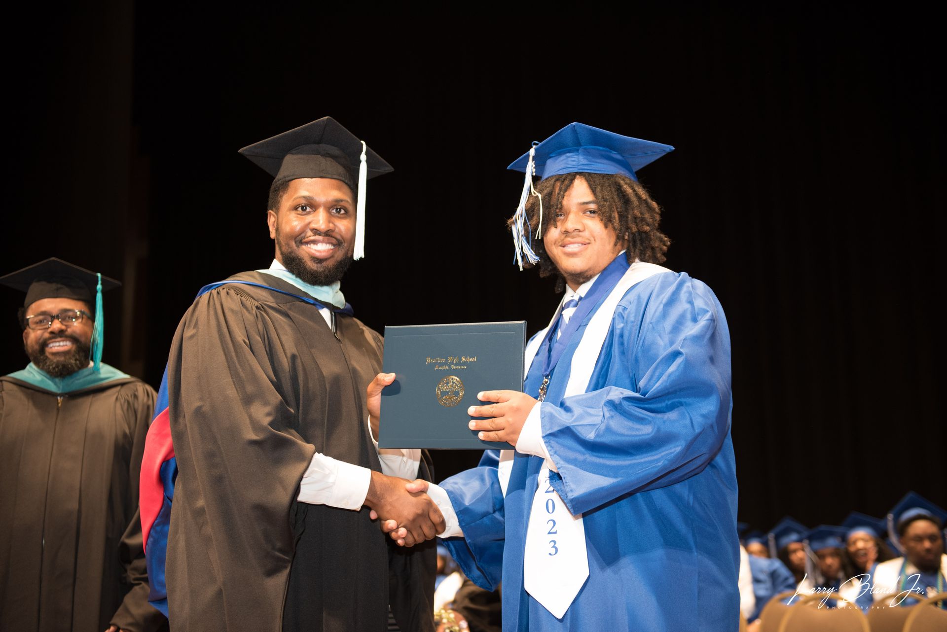 A graduate receives a diploma from a robed person on stage, with other graduates in the background.