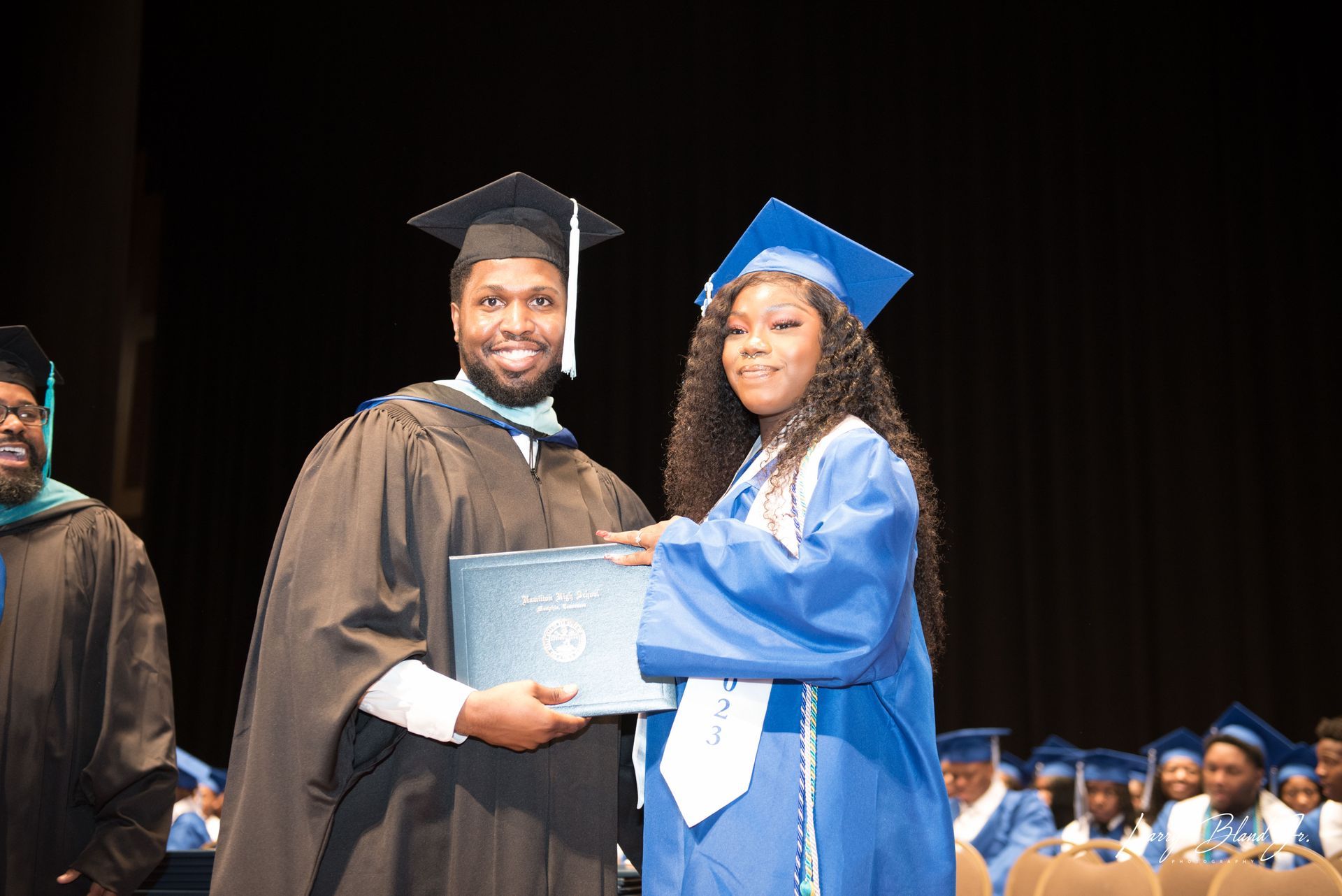 Graduation: Person in black gown hands a diploma to person in blue gown onstage.