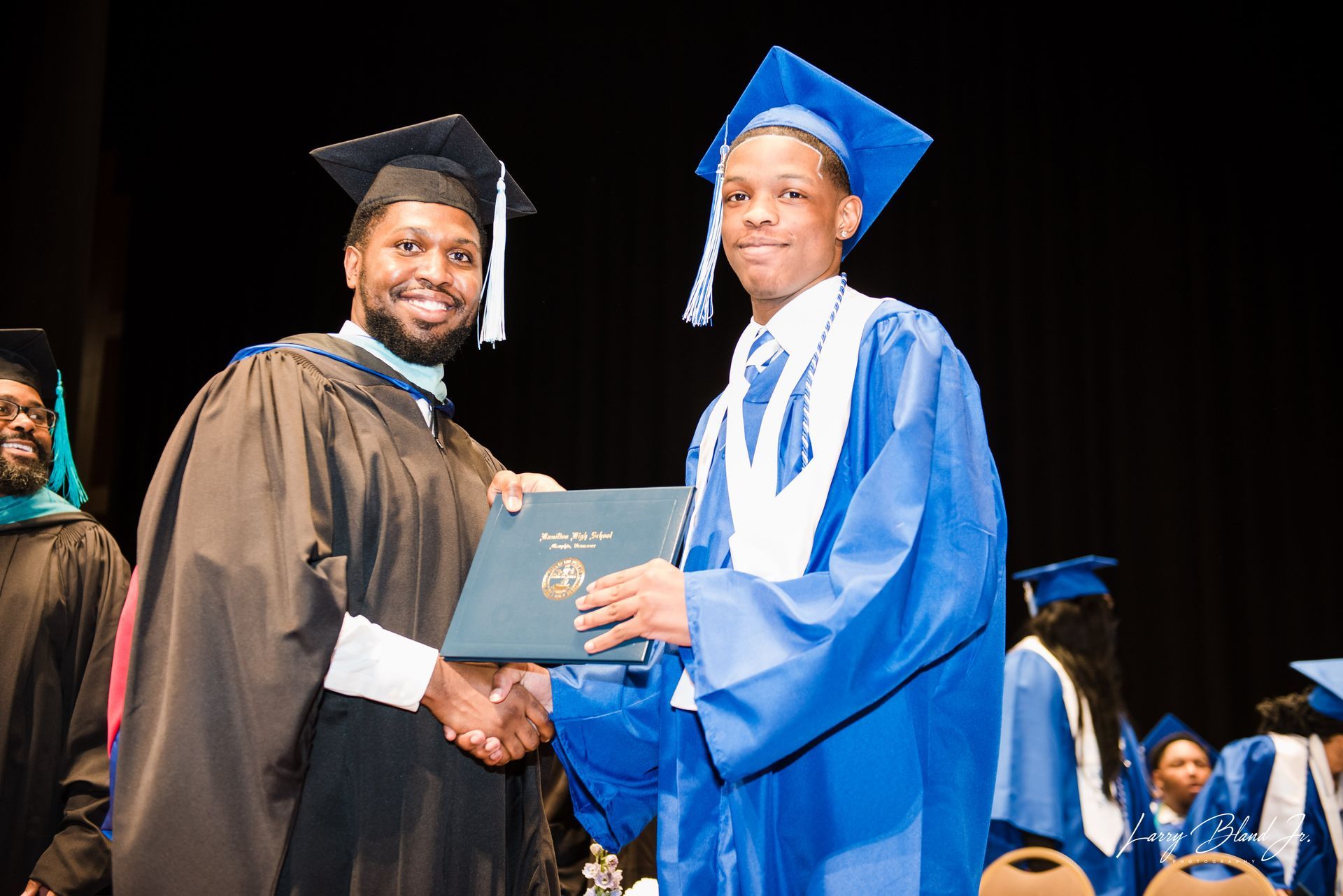 Man in academic robe hands diploma to graduate in blue gown.