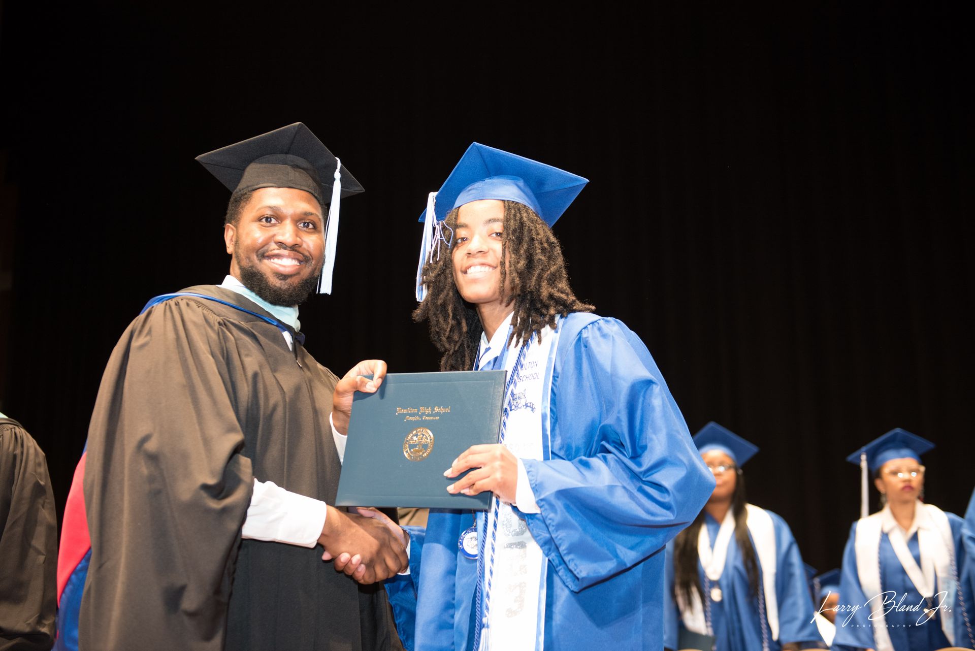 Man in academic robe hands diploma to smiling person in blue graduation gown.