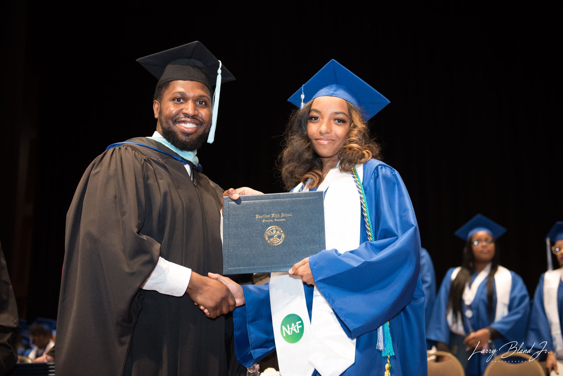 Person in black graduation gown hands diploma to person in blue gown at graduation.