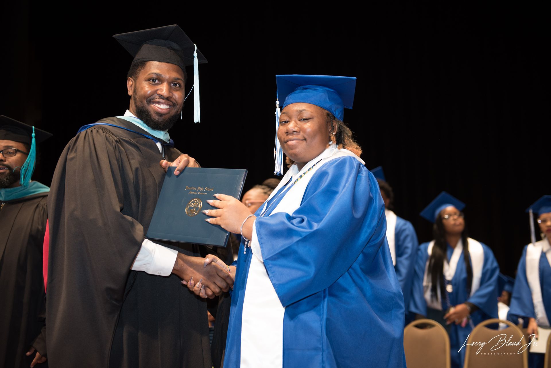 Man in black gown hands diploma to person in blue gown at graduation ceremony.