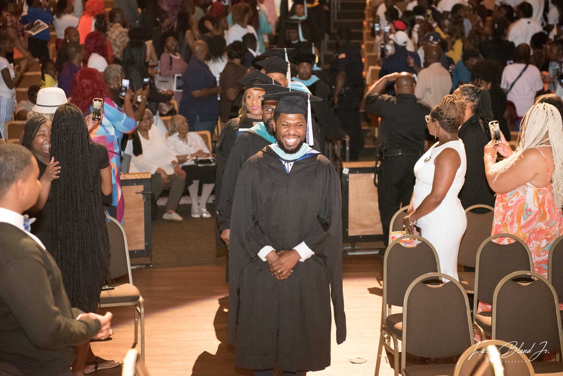 Graduates in black robes and mortarboards walk down an aisle during a ceremony. Spectators watch and take photos.