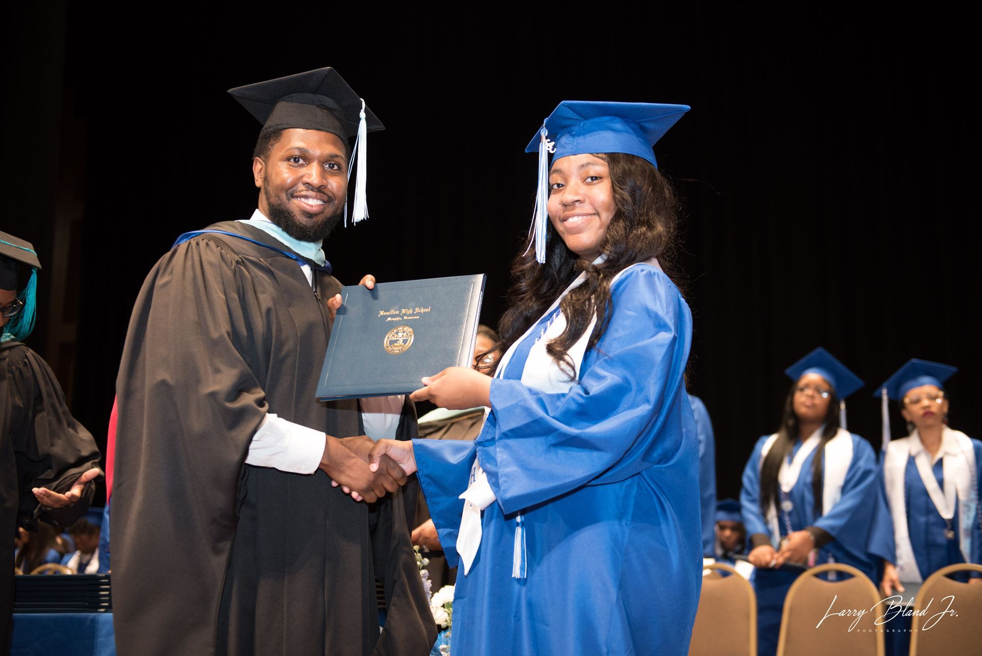 Graduation ceremony: Man in black gown hands diploma to person in blue gown; smiles exchanged.