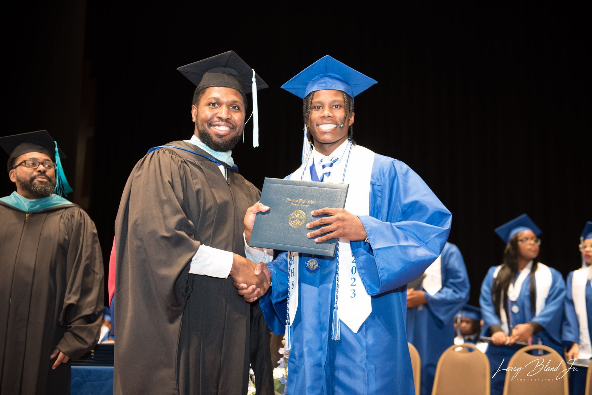 Man in black robe hands diploma to graduate in blue gown at ceremony.