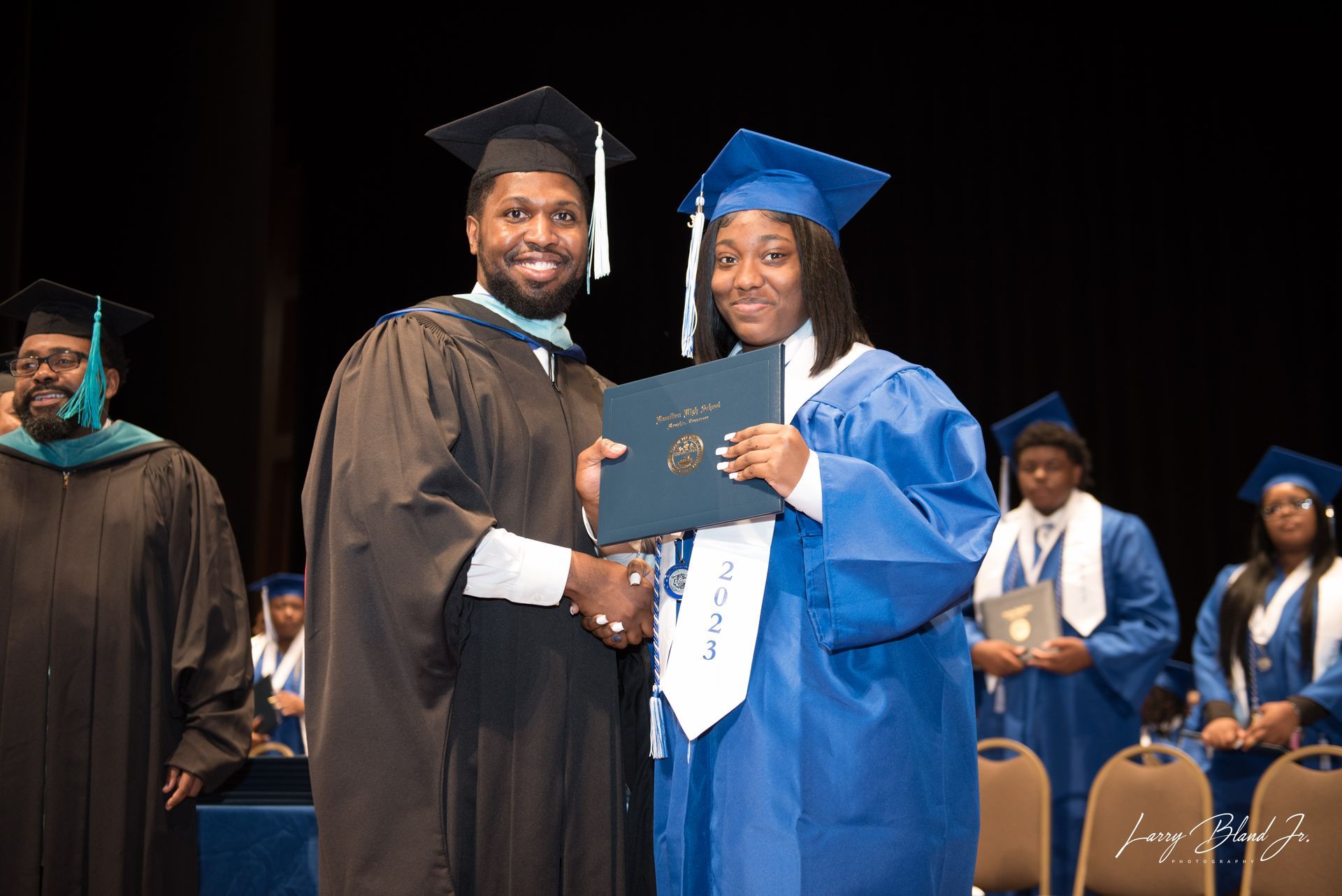 A graduate in blue robe receiving a diploma from a person in a black robe at a graduation ceremony.
