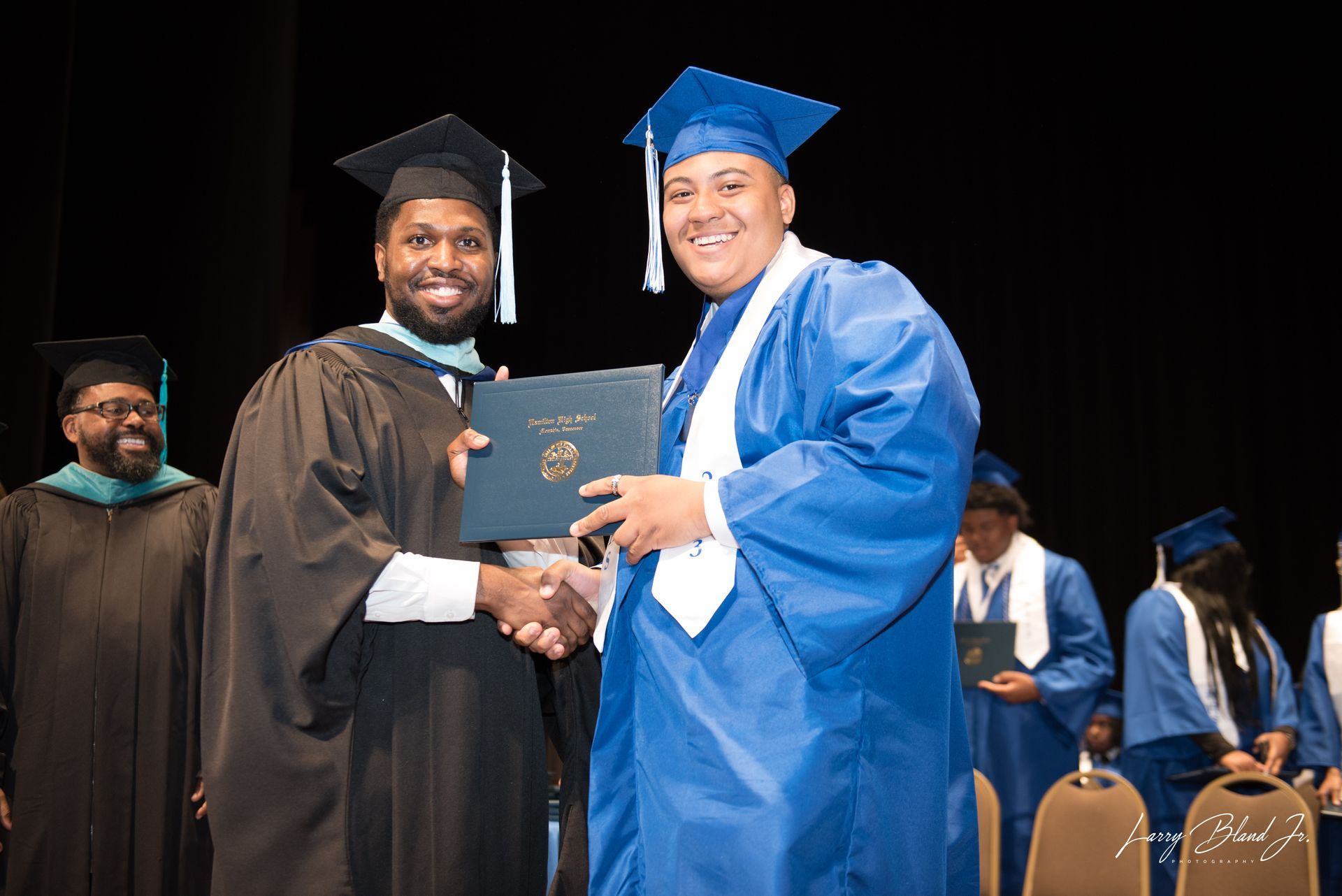 Two people in graduation gowns shaking hands on stage. The person on the right receives a diploma.