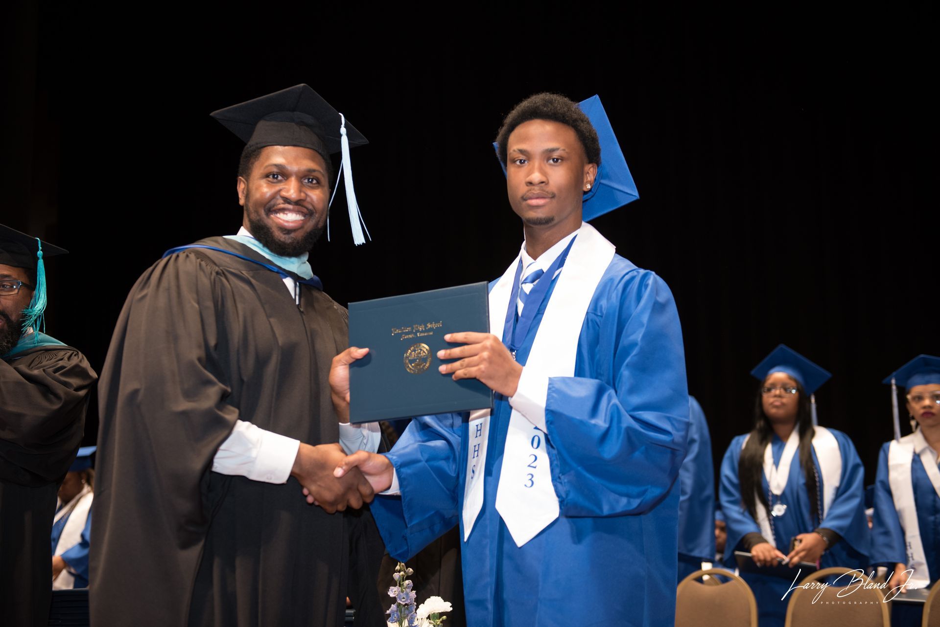 A person in academic regalia hands a diploma to a graduate wearing a blue gown at a ceremony.