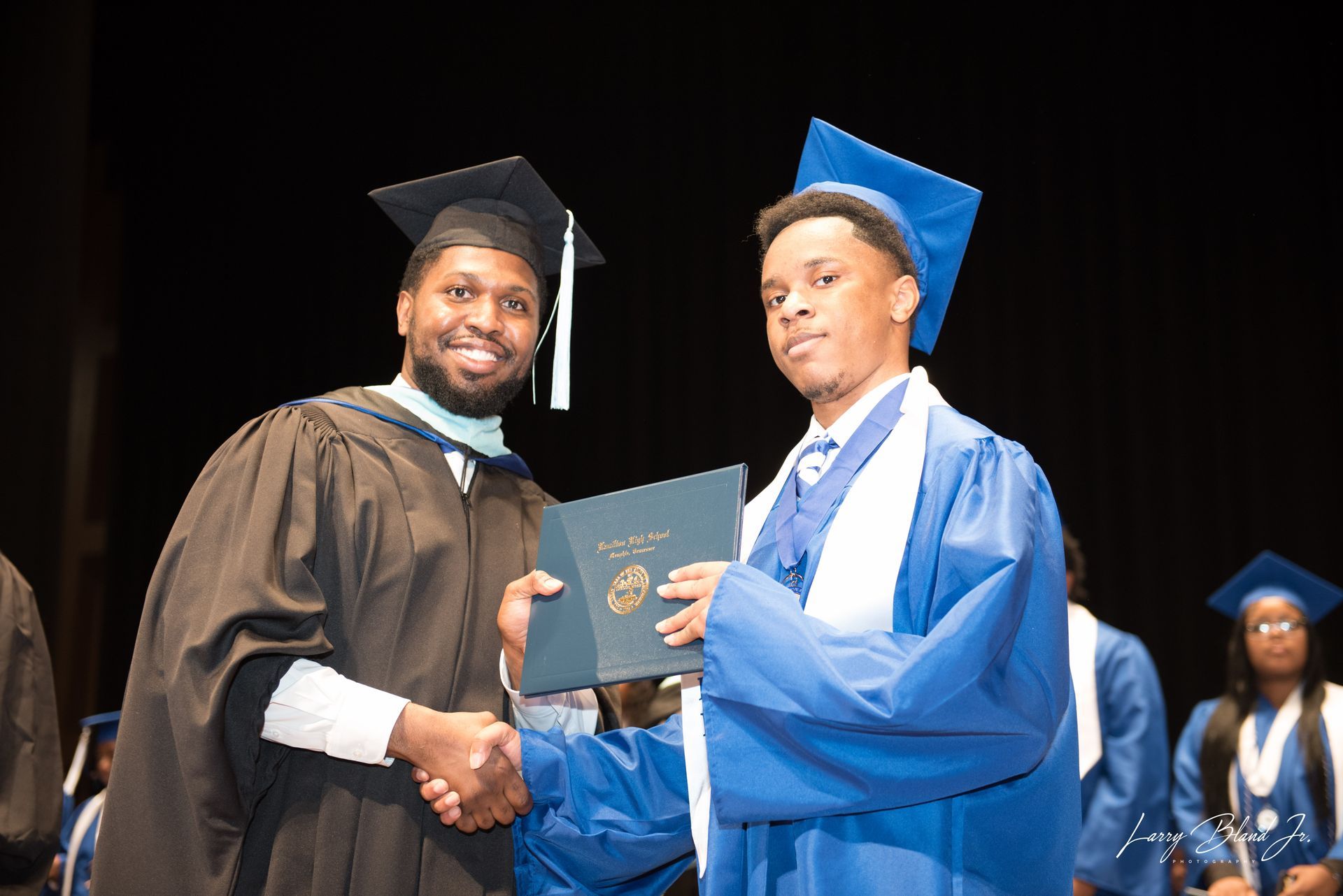A person in black graduation attire hands a diploma to a person in blue graduation attire.