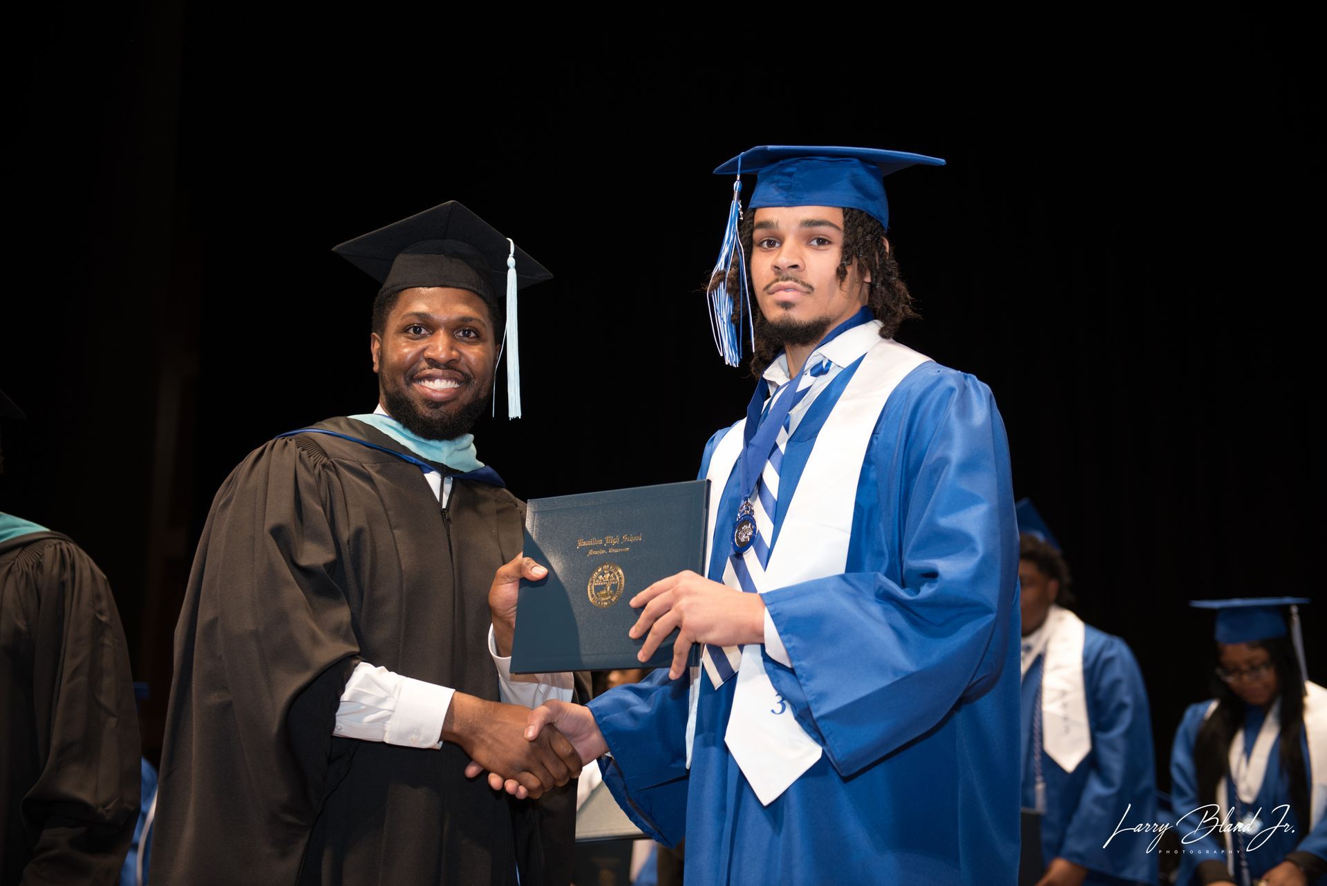 Man in black robe shakes hands with a graduate in blue gown, holding a diploma onstage.