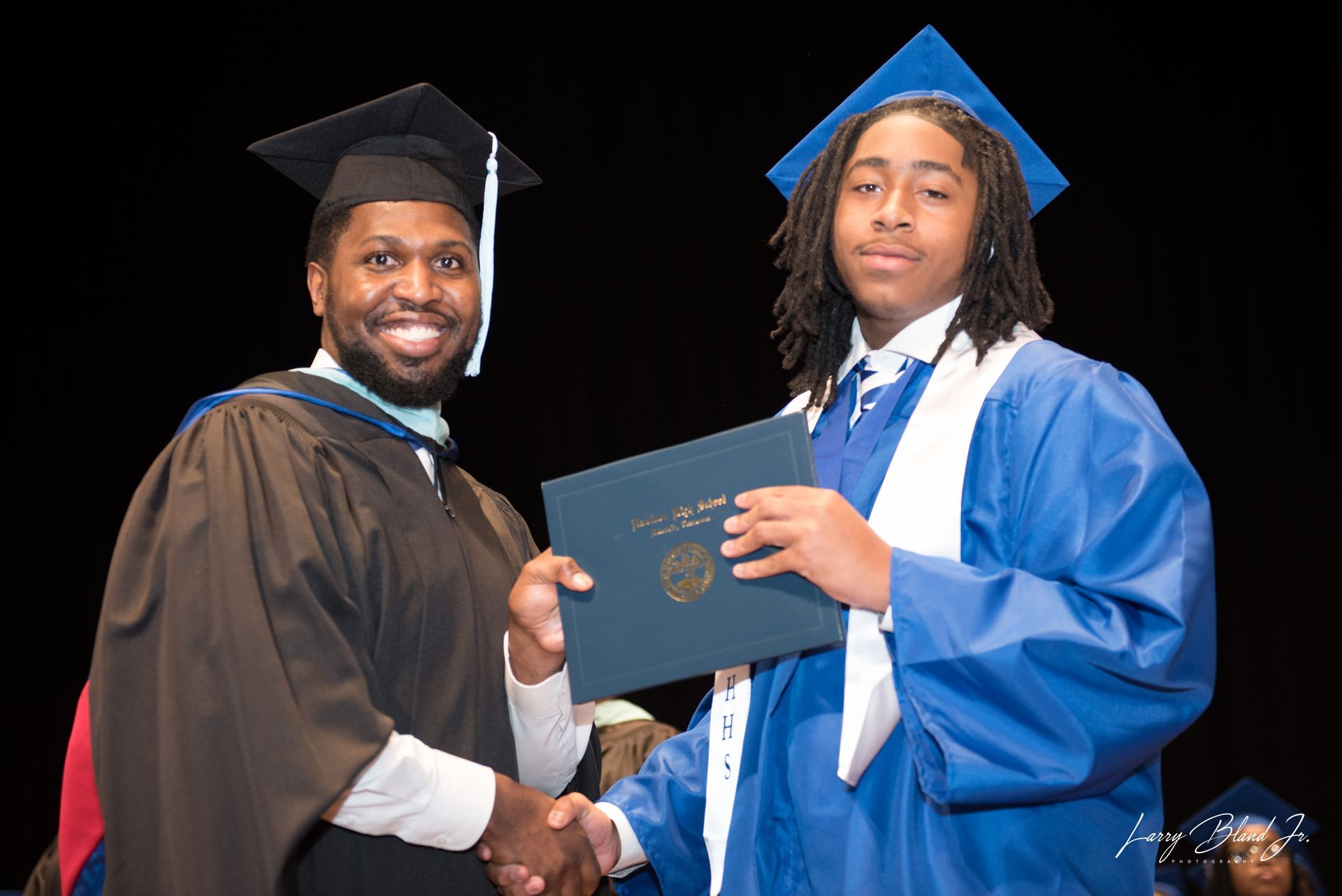 Person in graduation gown shaking hands with a graduate in blue gown, holding diploma. Stage setting.