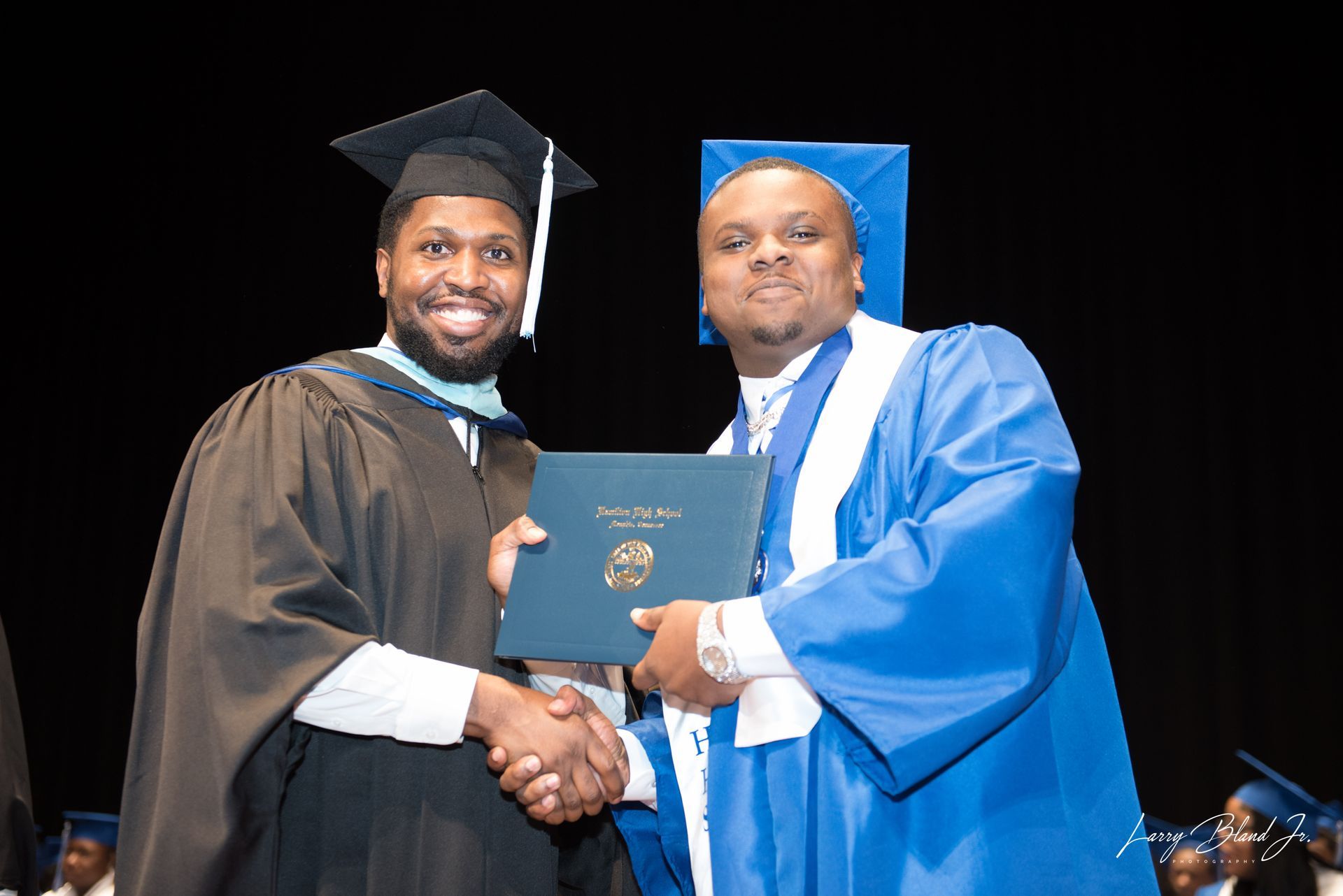 Man in graduation gown receiving diploma and shaking hands with another man in a blue gown onstage.