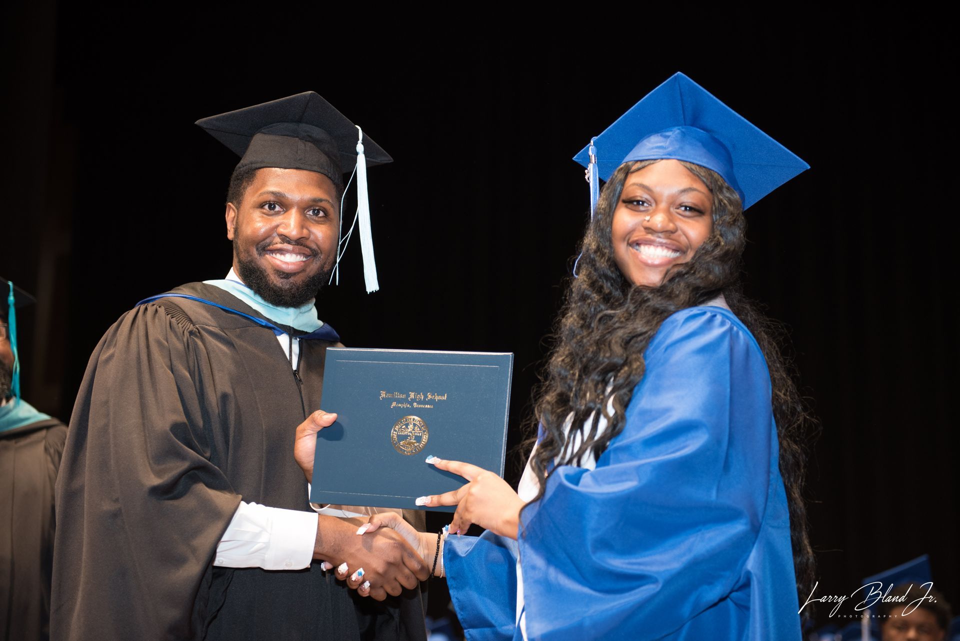 Man in dark gown shakes hands with person in blue cap and gown, handing over a diploma.