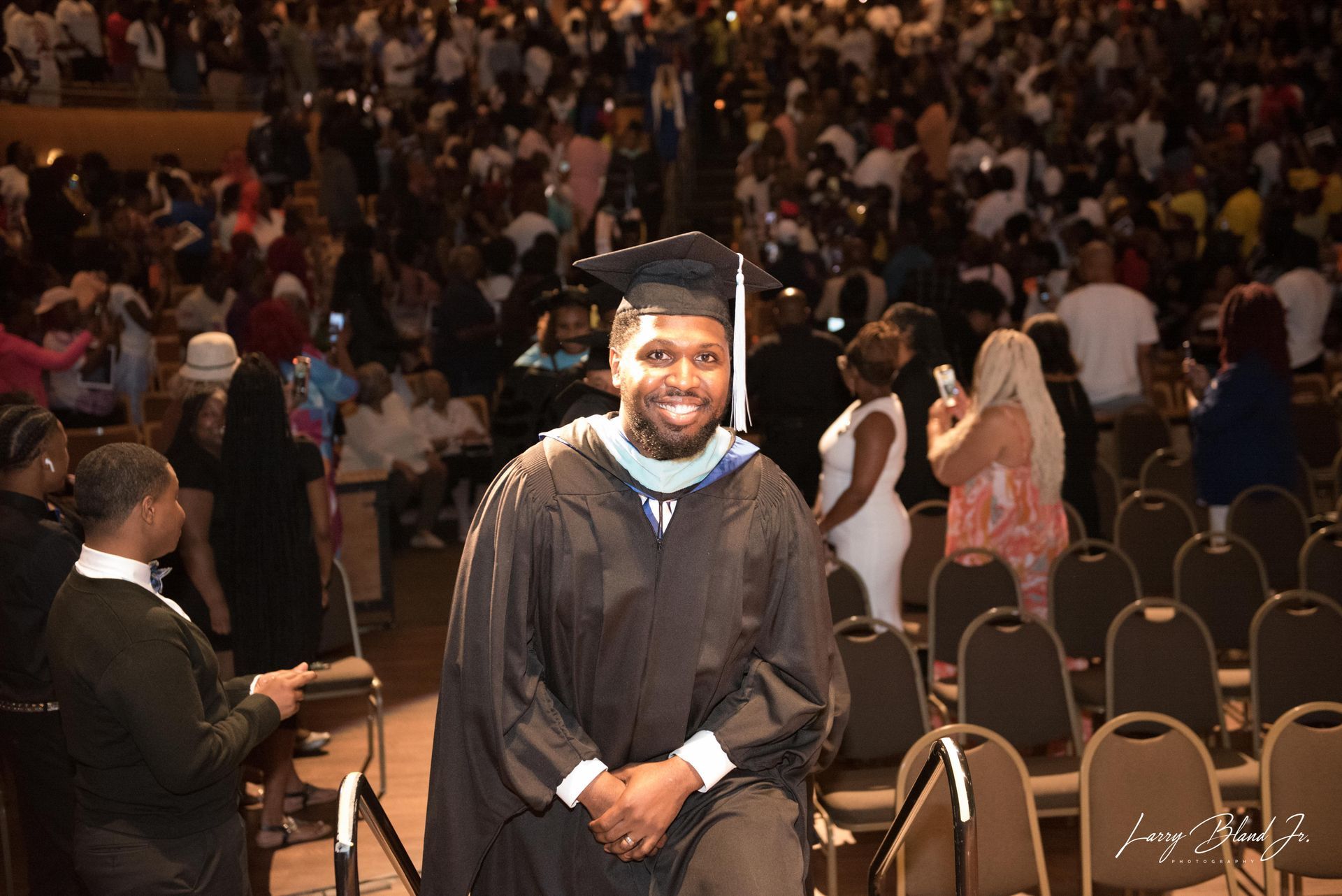 Smiling graduate in cap and gown, standing in an auditorium full of people.