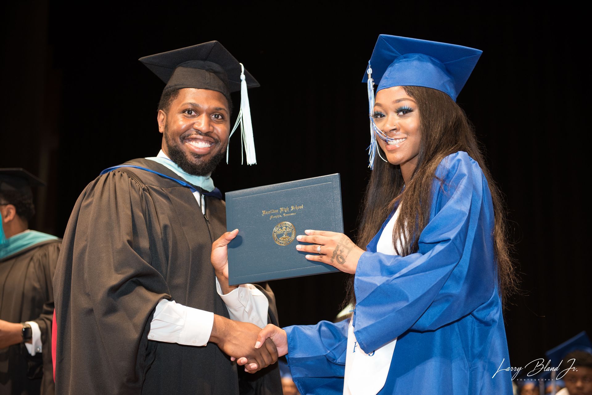 Man in black robe hands diploma to woman in blue gown; both shake hands.