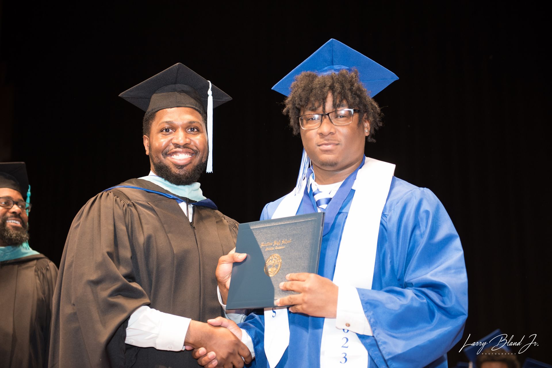 Man in black graduation robes shakes hands with a man in a blue gown, presenting a diploma on stage.