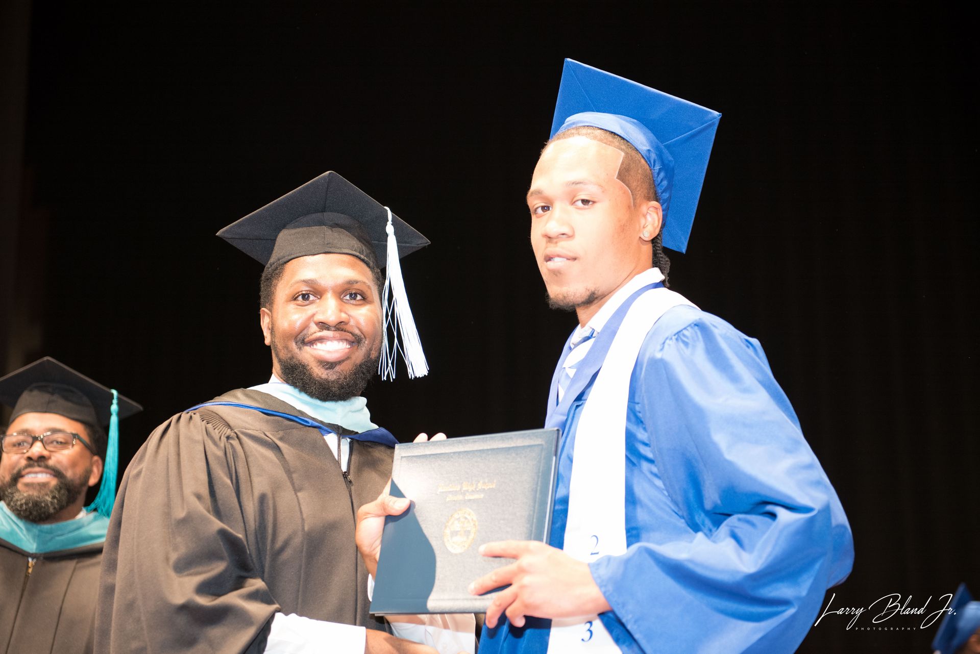 A graduate in blue robe receiving diploma from a person in black robe with a smile on stage.