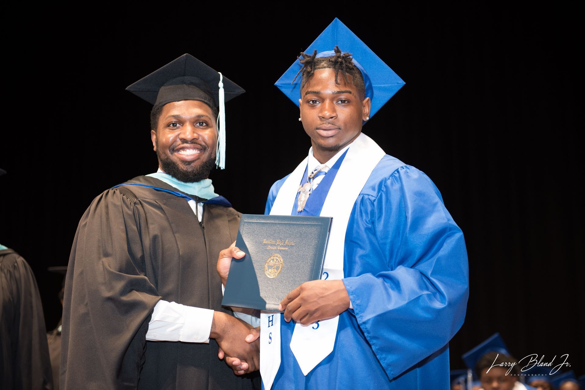 Man in black gown shakes hands with person in blue graduation attire, diploma in hand.