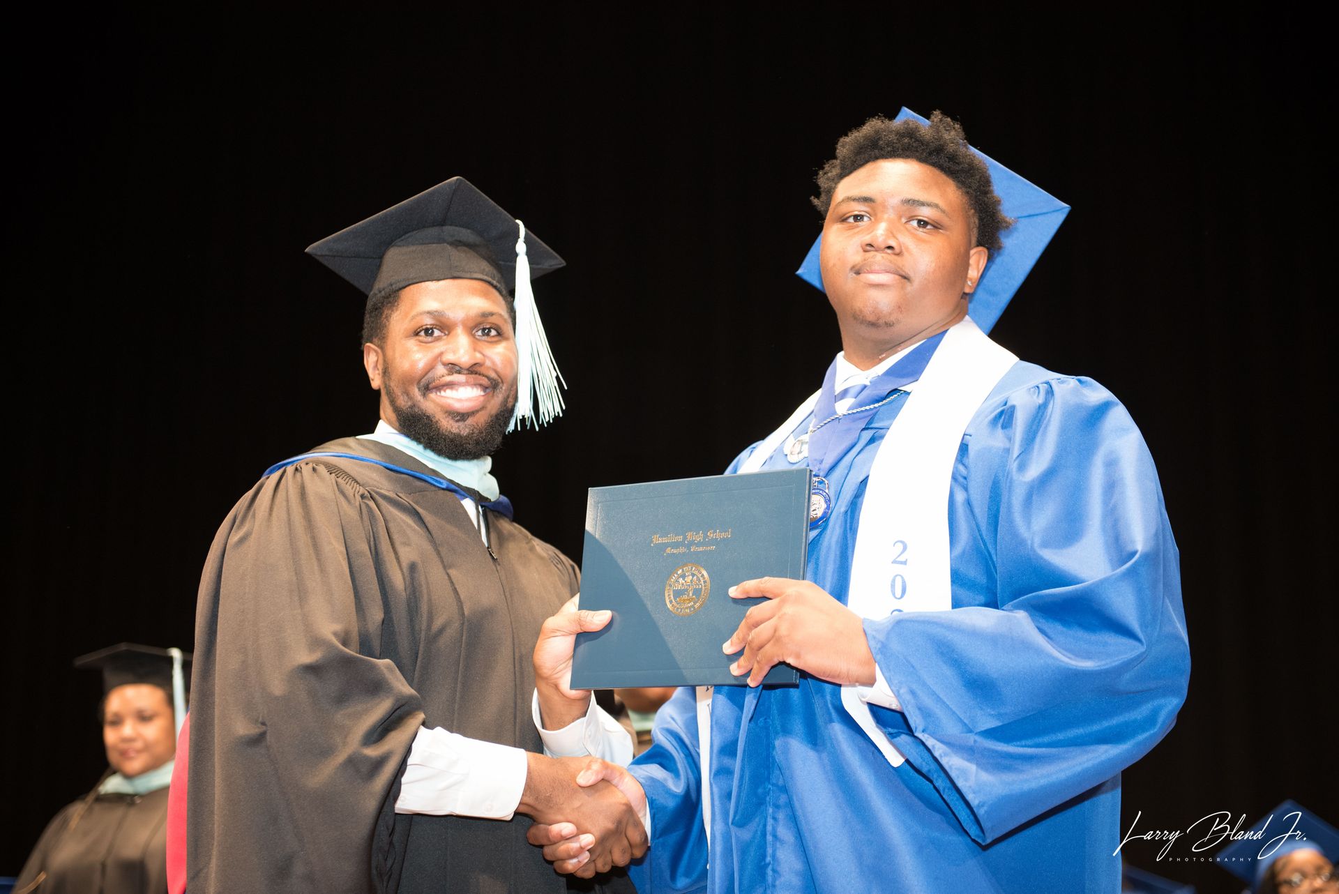 Man in blue graduation gown receives diploma from man in brown academic regalia.