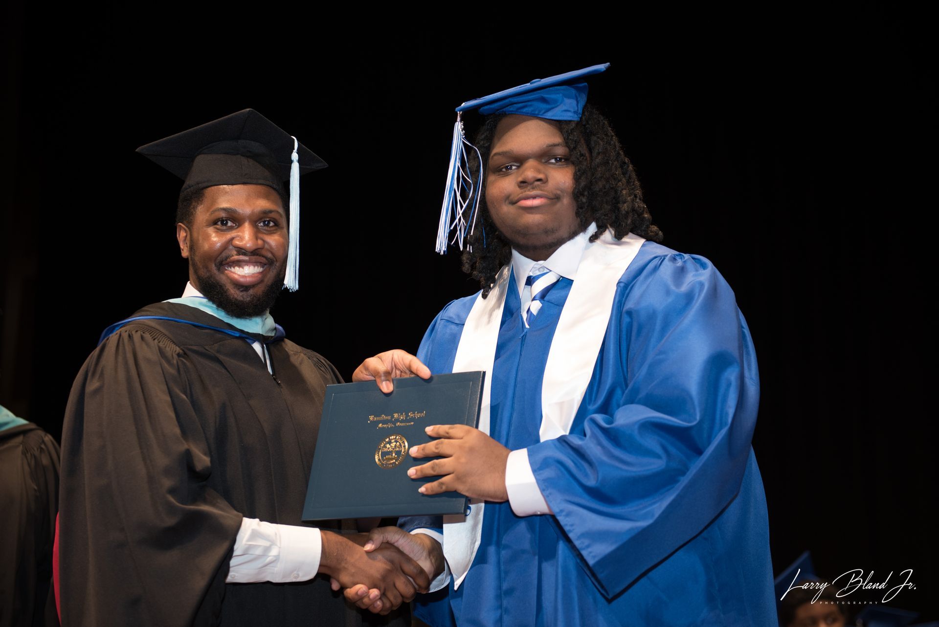 Graduation: Man in black robe hands diploma to graduate in blue gown; they shake hands.