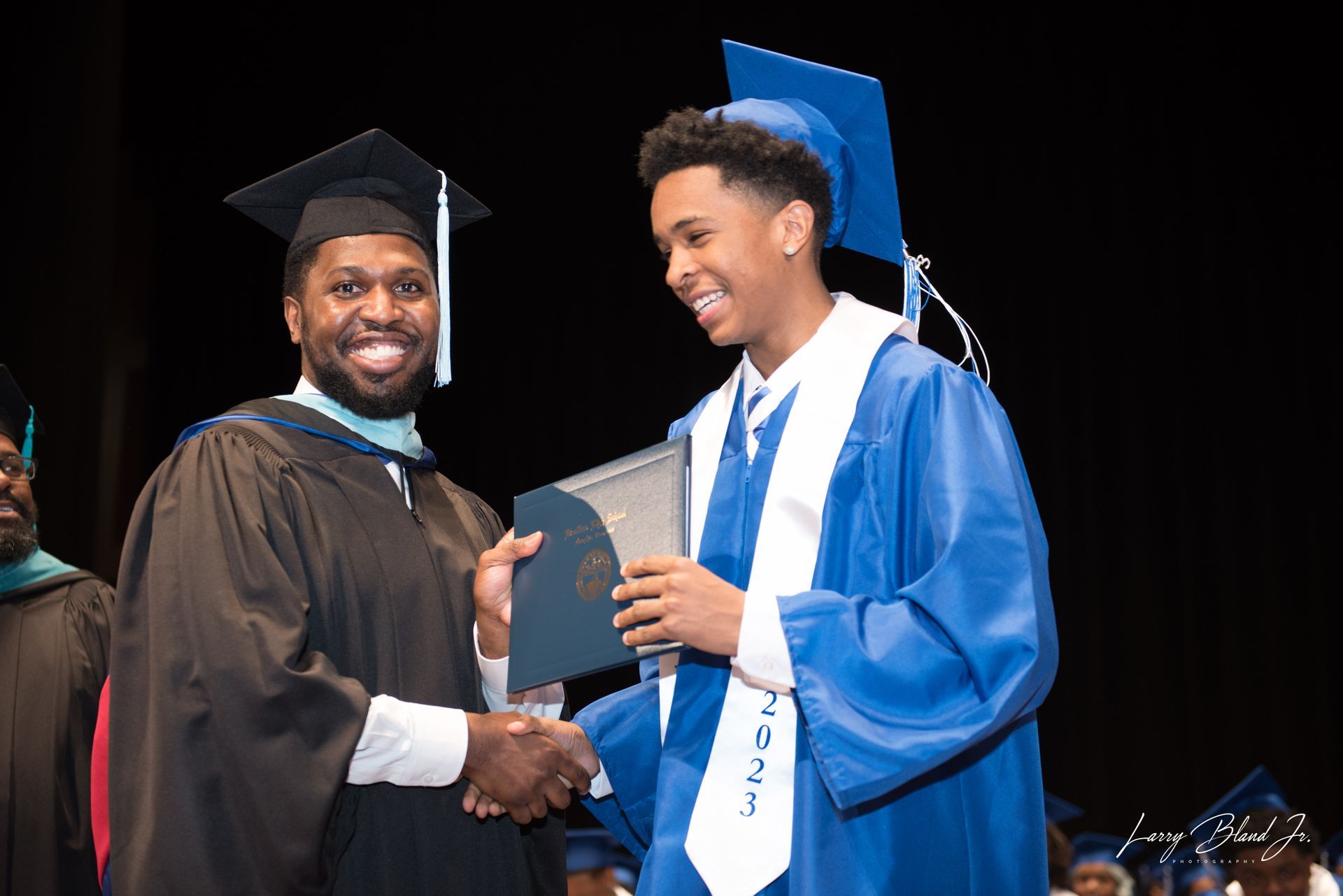 Man in cap and gown shakes hands with a graduate receiving a diploma, both smiling. Blue and black robes.