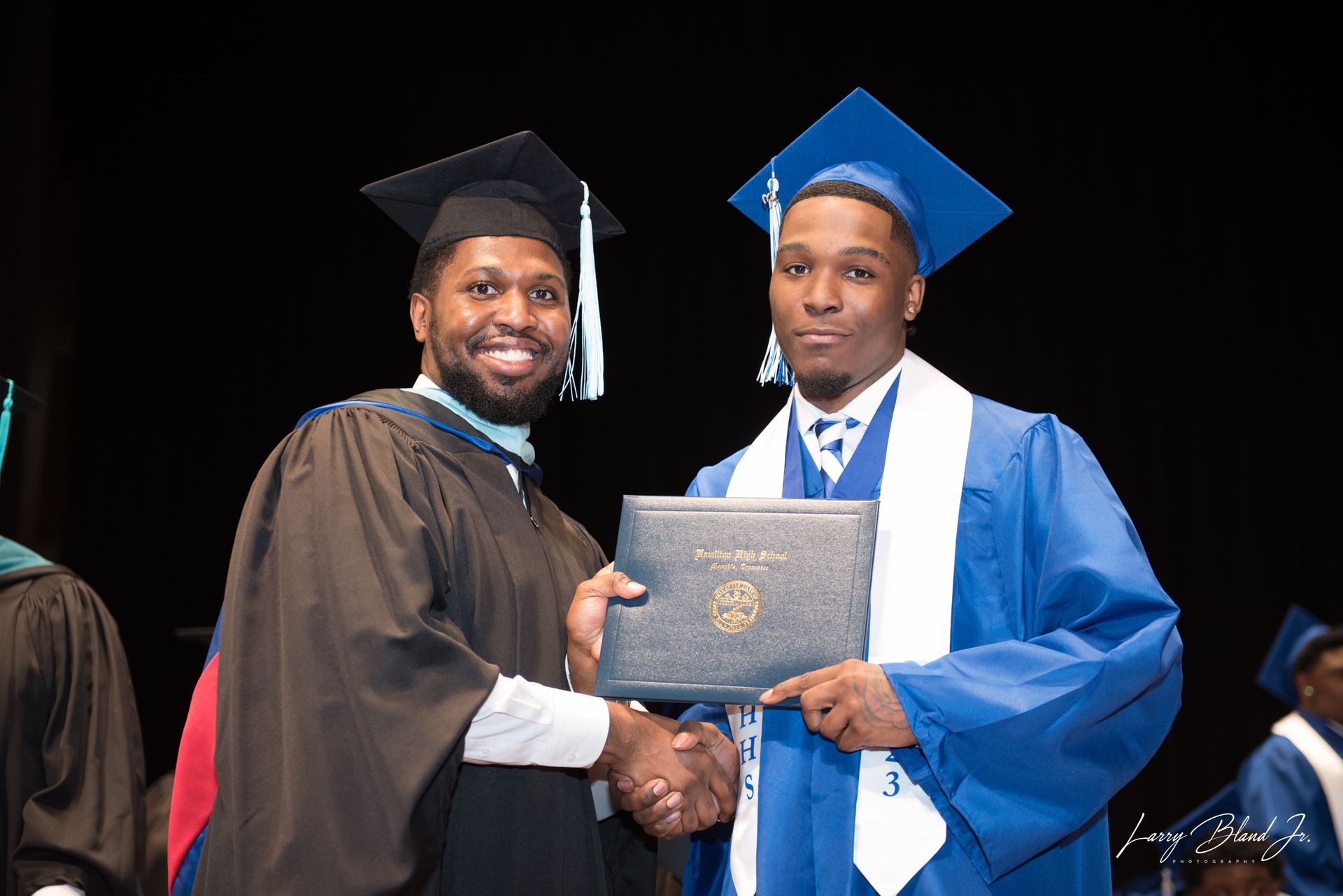 Man in cap and gown presents diploma to graduate in blue gown, shaking hands at a ceremony.
