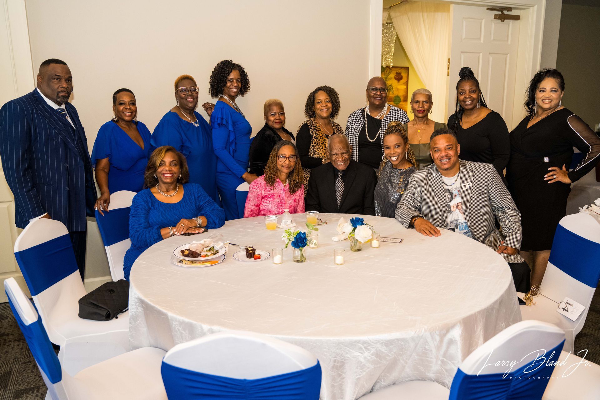 Group of people gathered around a decorated table, many wearing blue. Indoor setting, white walls.