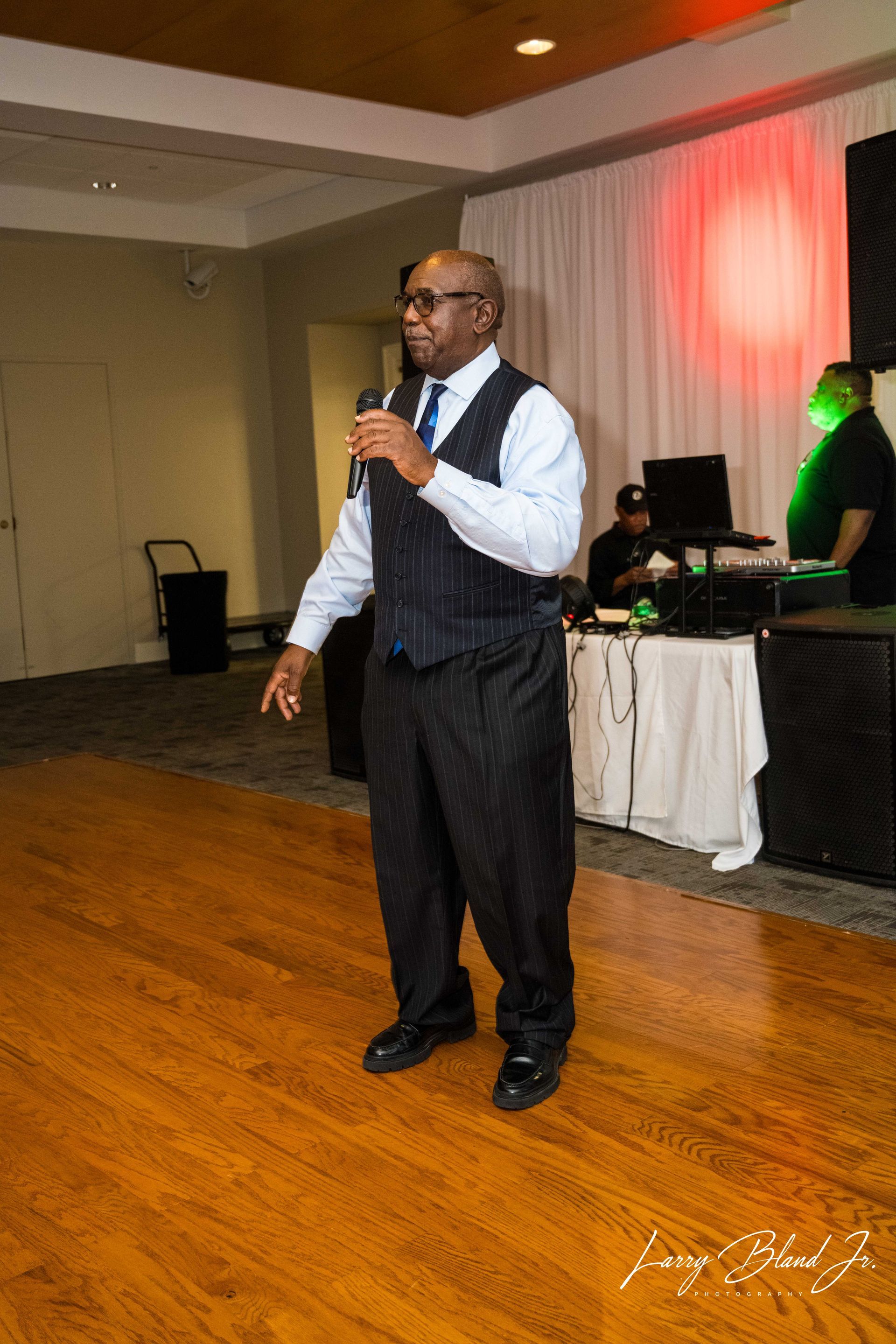 Man in formal wear speaking into a microphone at an event with DJ setup in background.