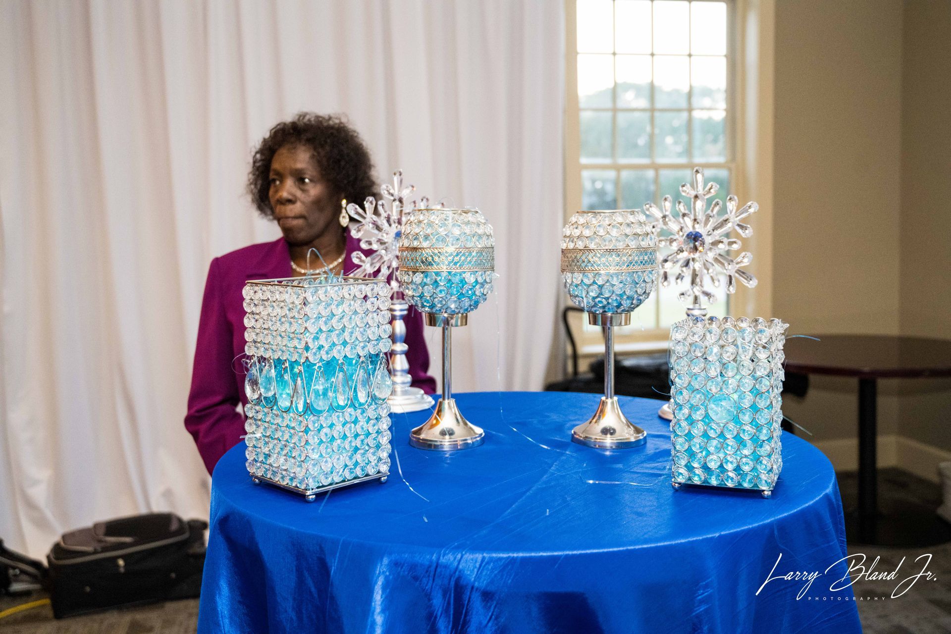 Woman in magenta jacket stands by a blue table with decorative crystal and teal centerpieces.