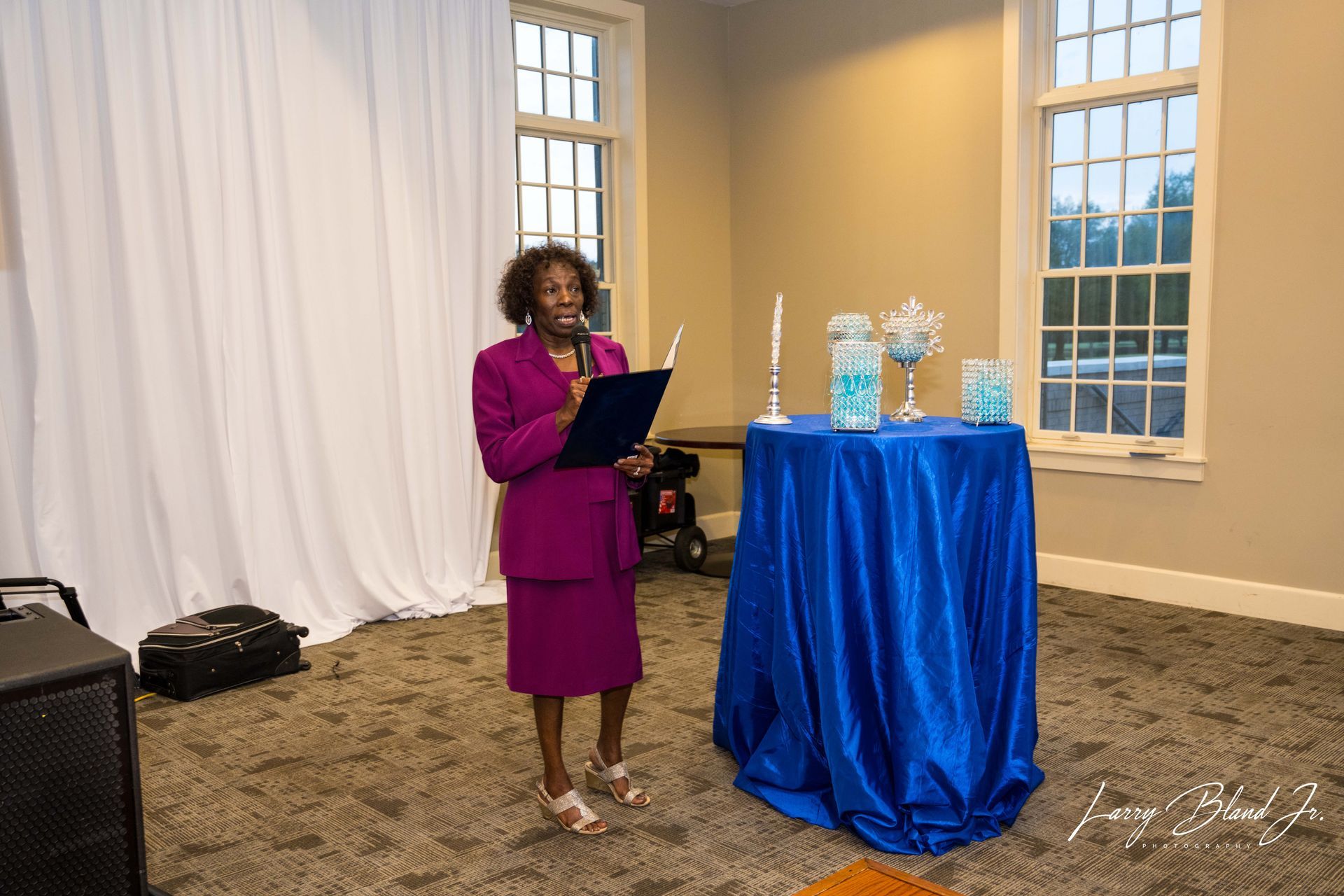 Woman in magenta suit speaking at podium by a blue-covered table with candles and vases.