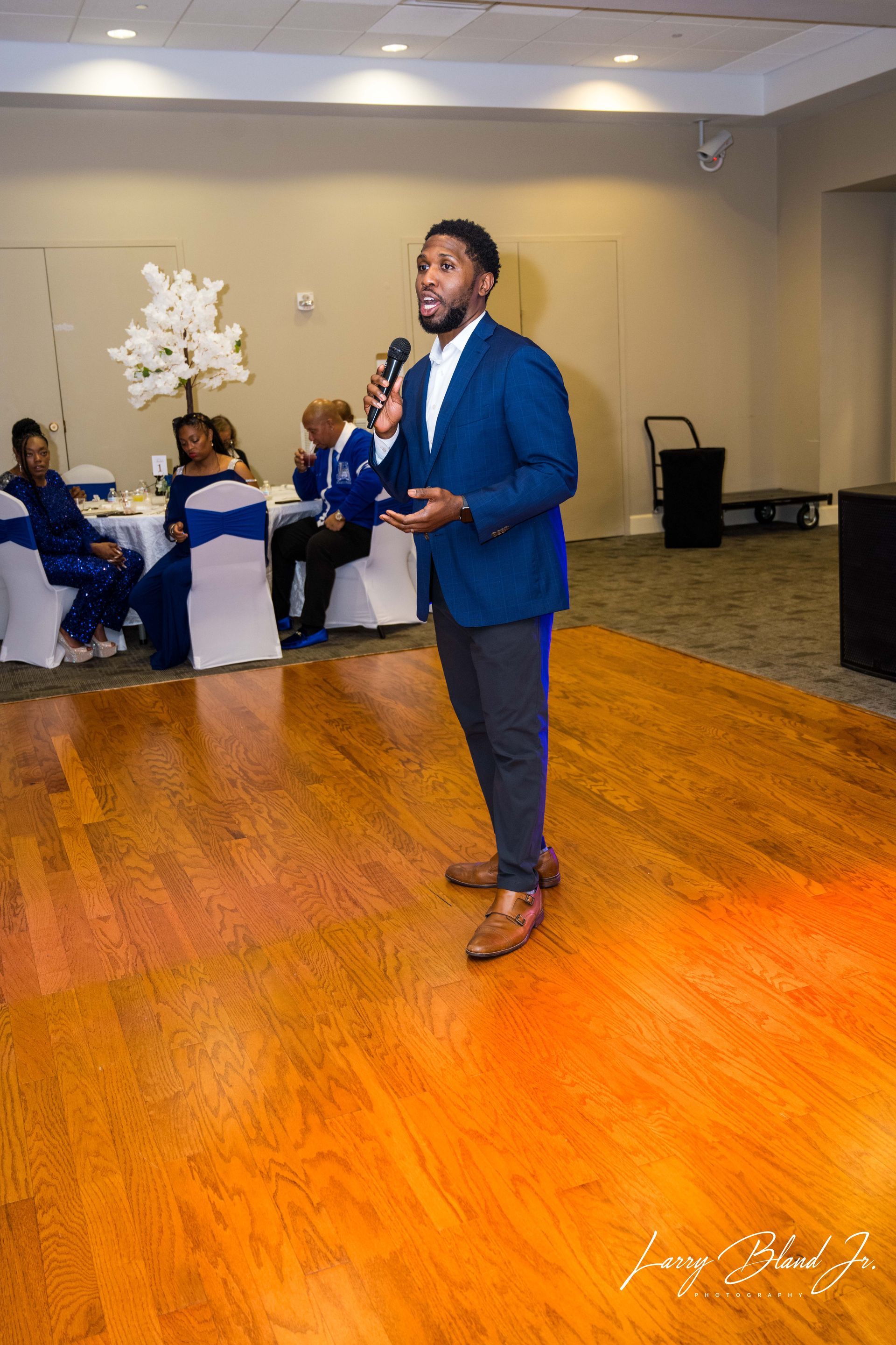 Man in blue blazer speaks into microphone at event, wooden floor, seated guests in background.