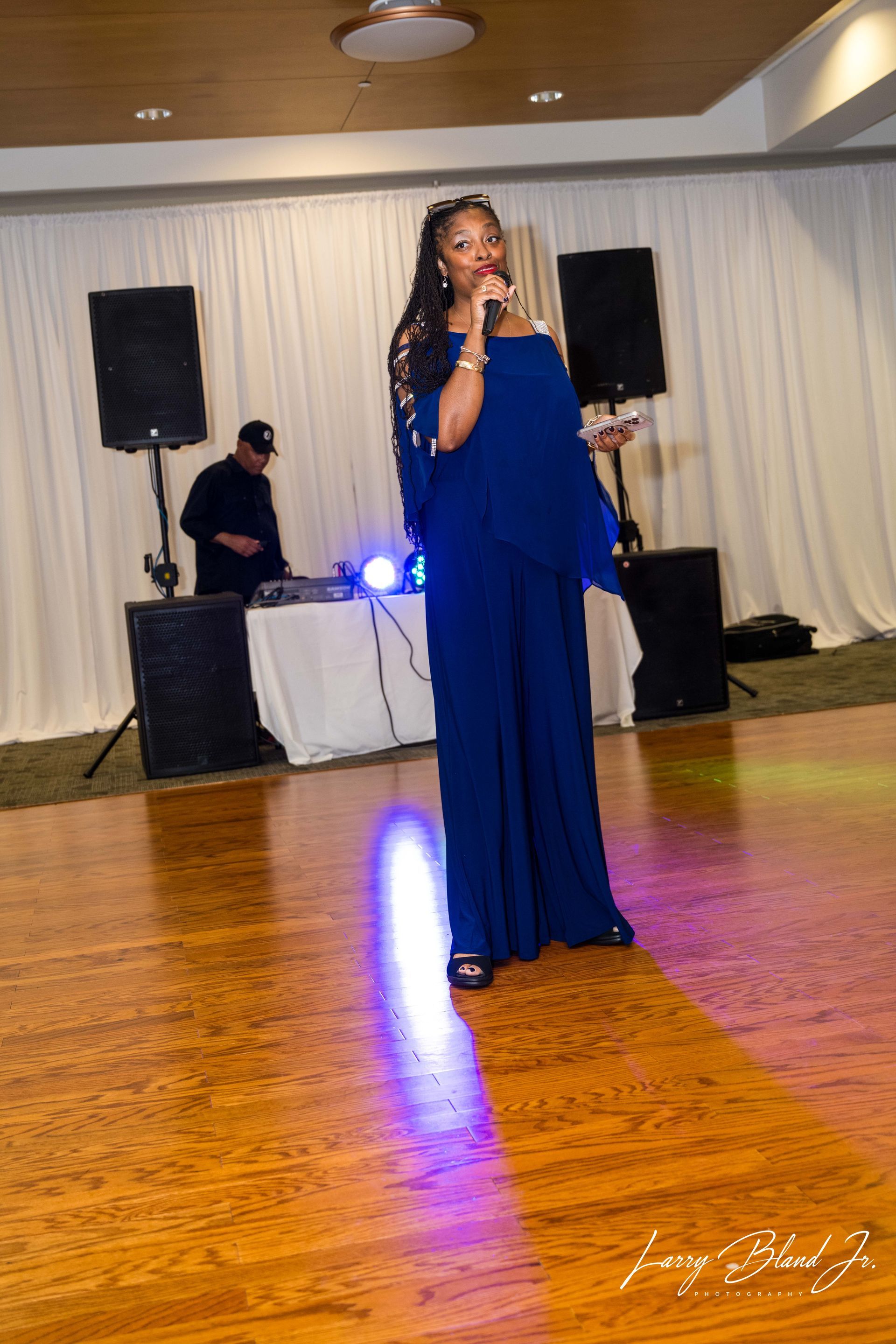 Woman in blue jumpsuit speaking into a microphone on a dance floor, DJ in background.
