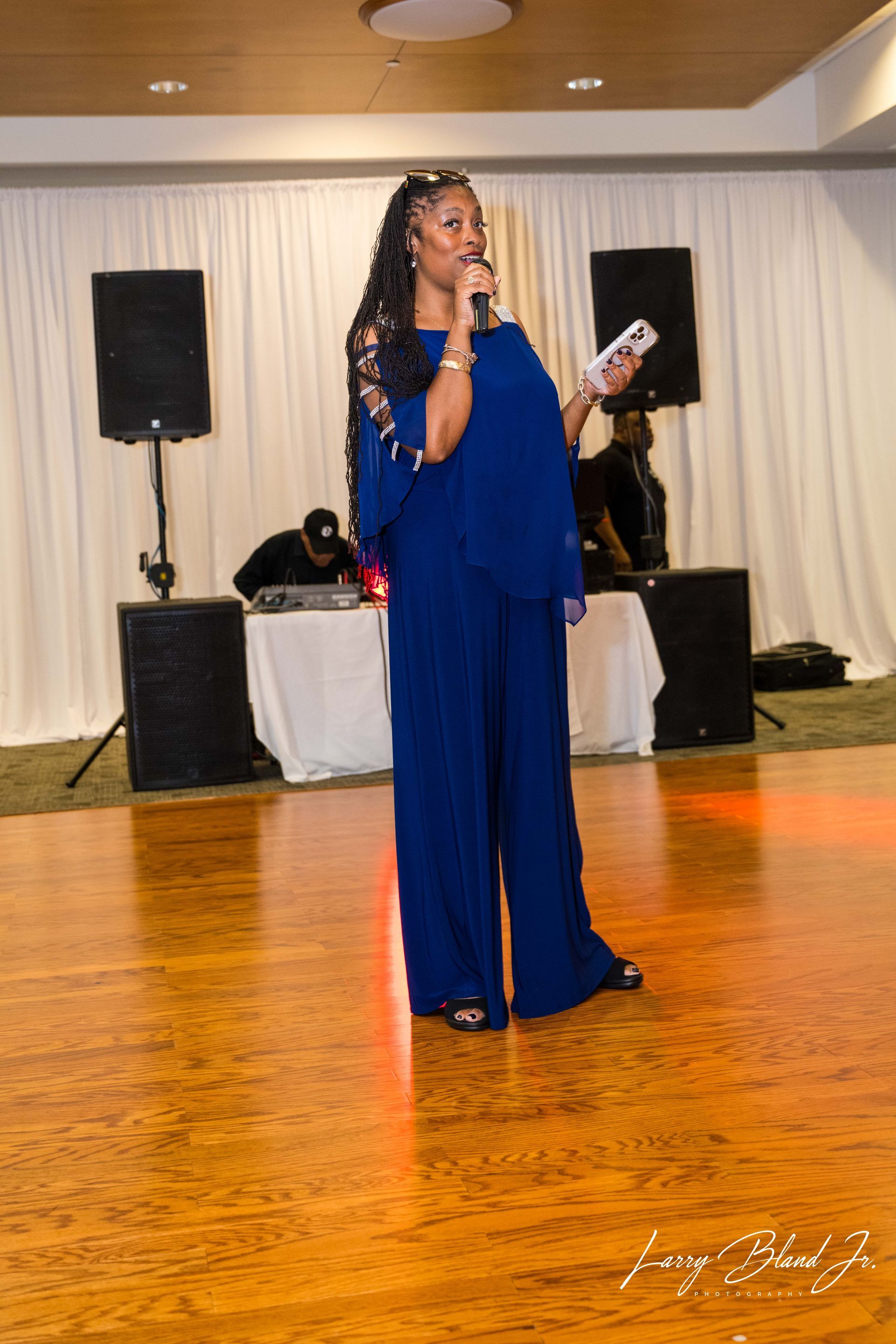 Woman in blue jumpsuit speaking into microphone on a dance floor, DJ in the background.