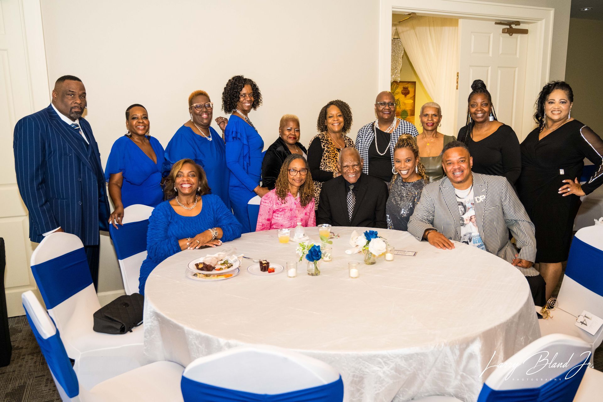 Group of people gathered around a white table. Many wear blue attire, smiling. Indoors, festive setting.