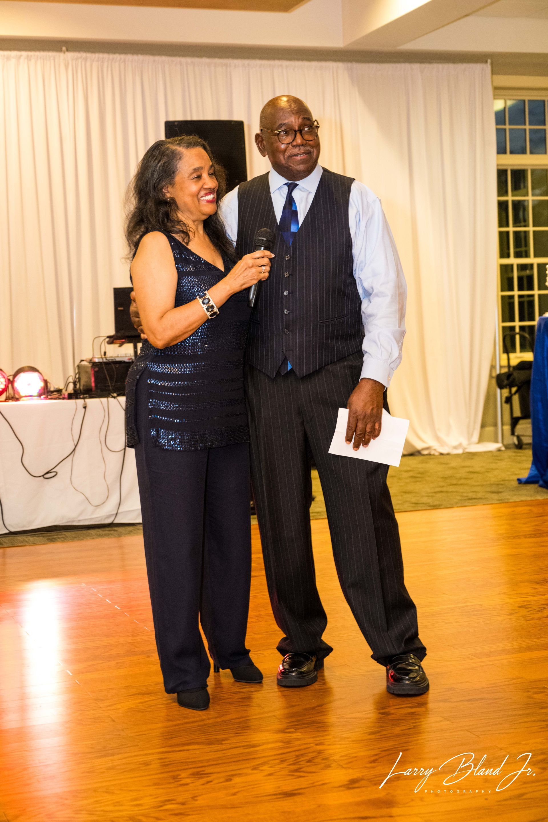 Two people smiling, holding a microphone, on a dance floor. Formal attire, with a background of curtains and windows.