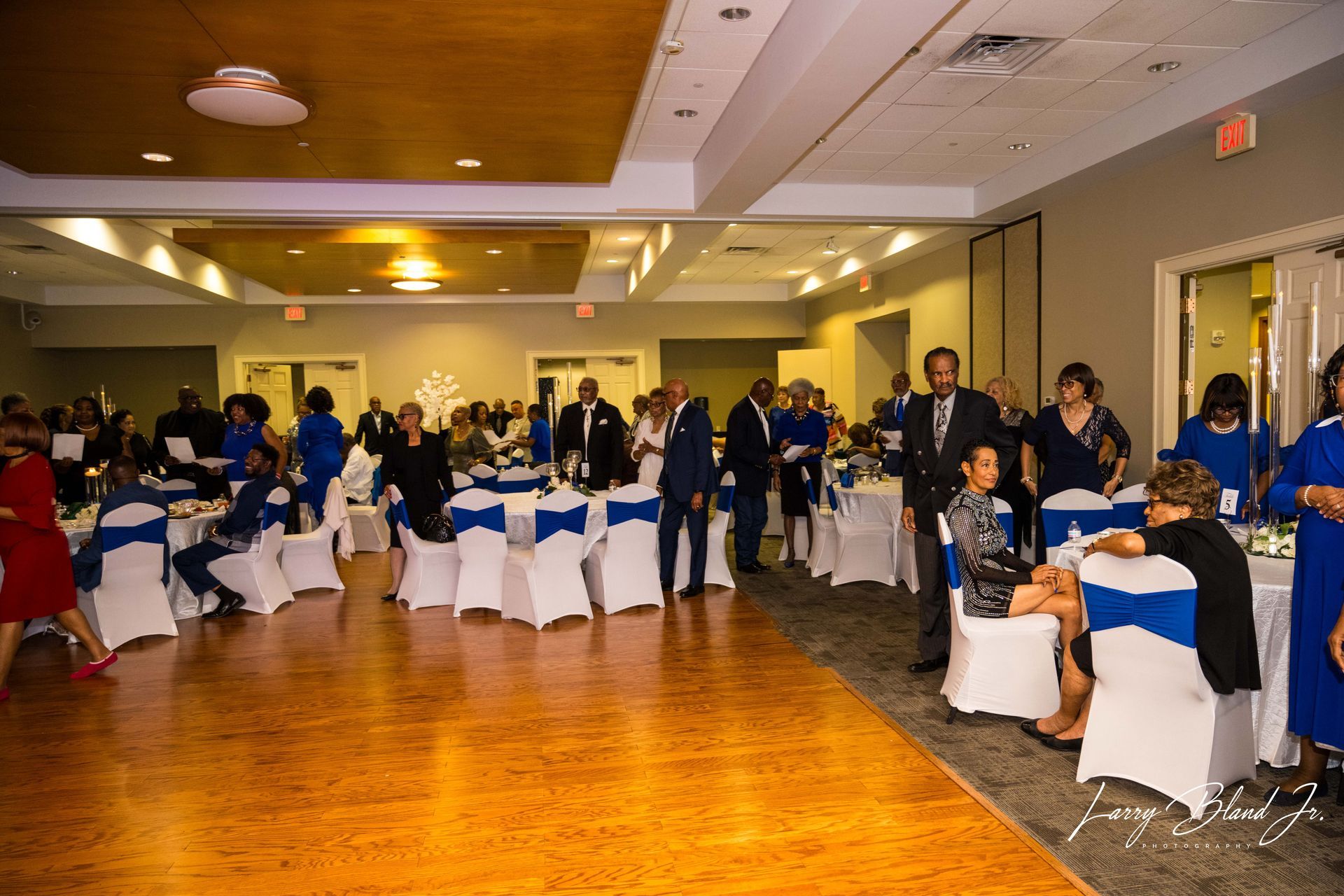 People at a formal event in a ballroom with tables, chairs, and a dance floor; blue and white color scheme.