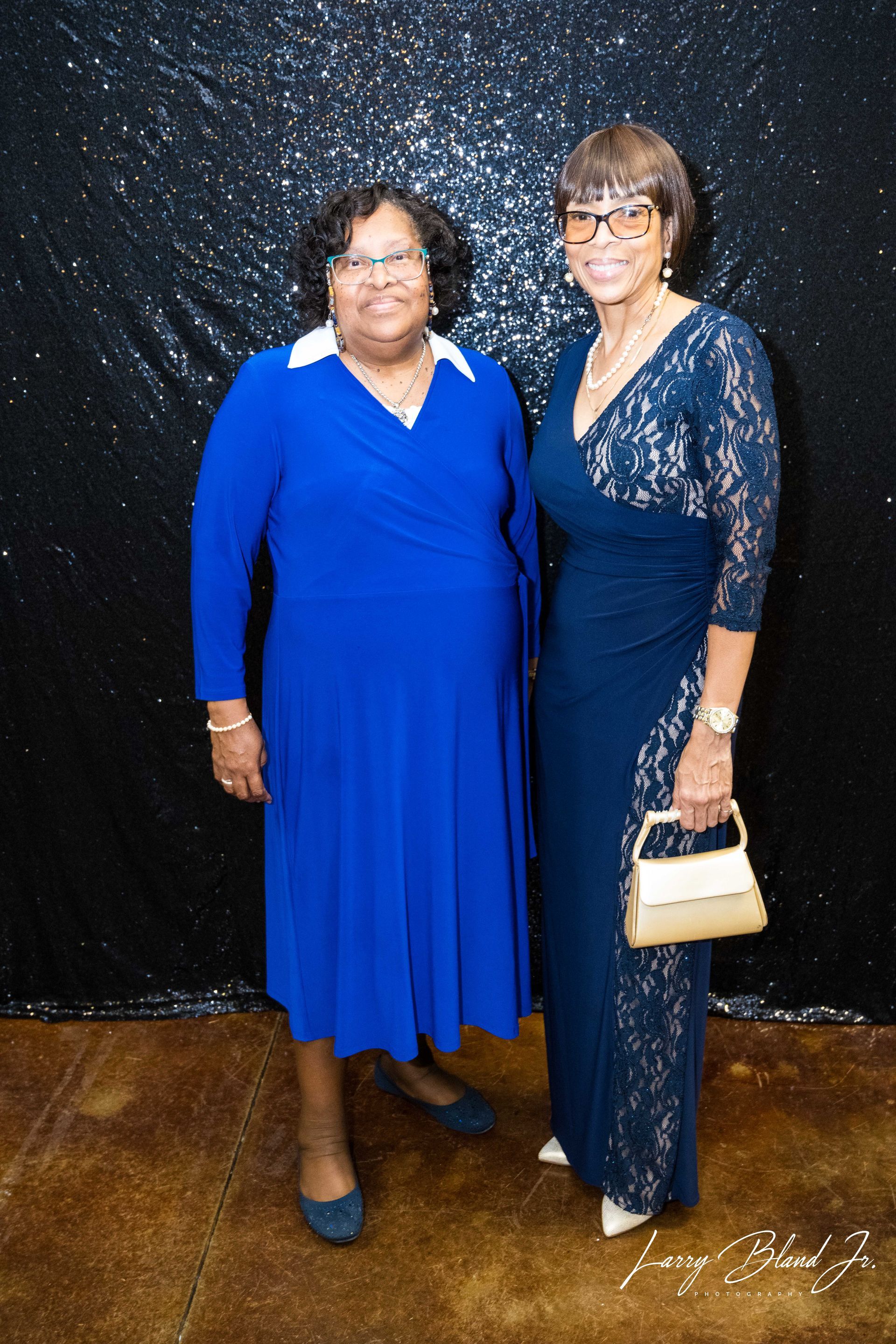 Two women in formal attire pose in front of a sparkly black backdrop.