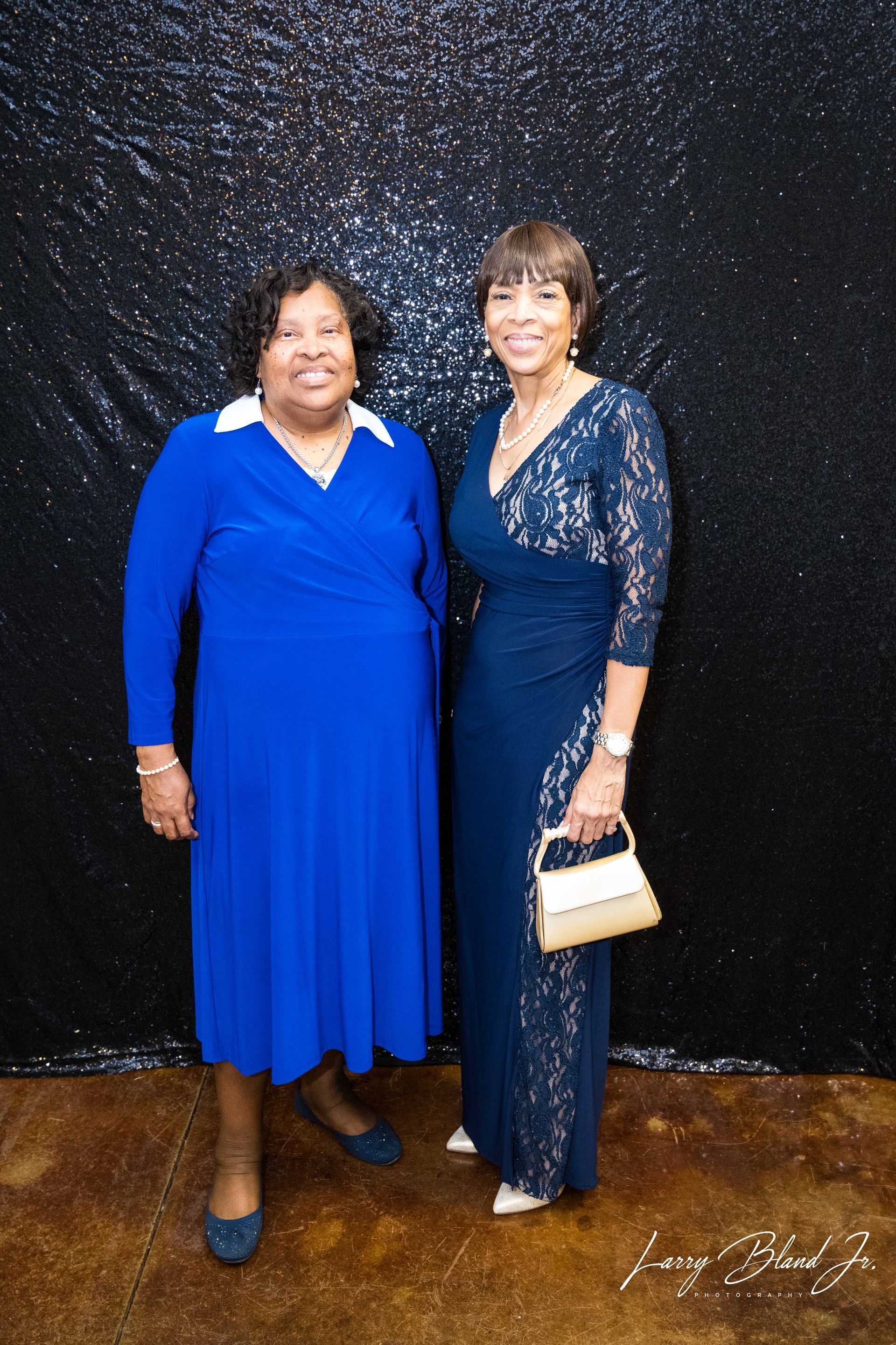 Two women in formal attire pose in front of a sparkly black backdrop.