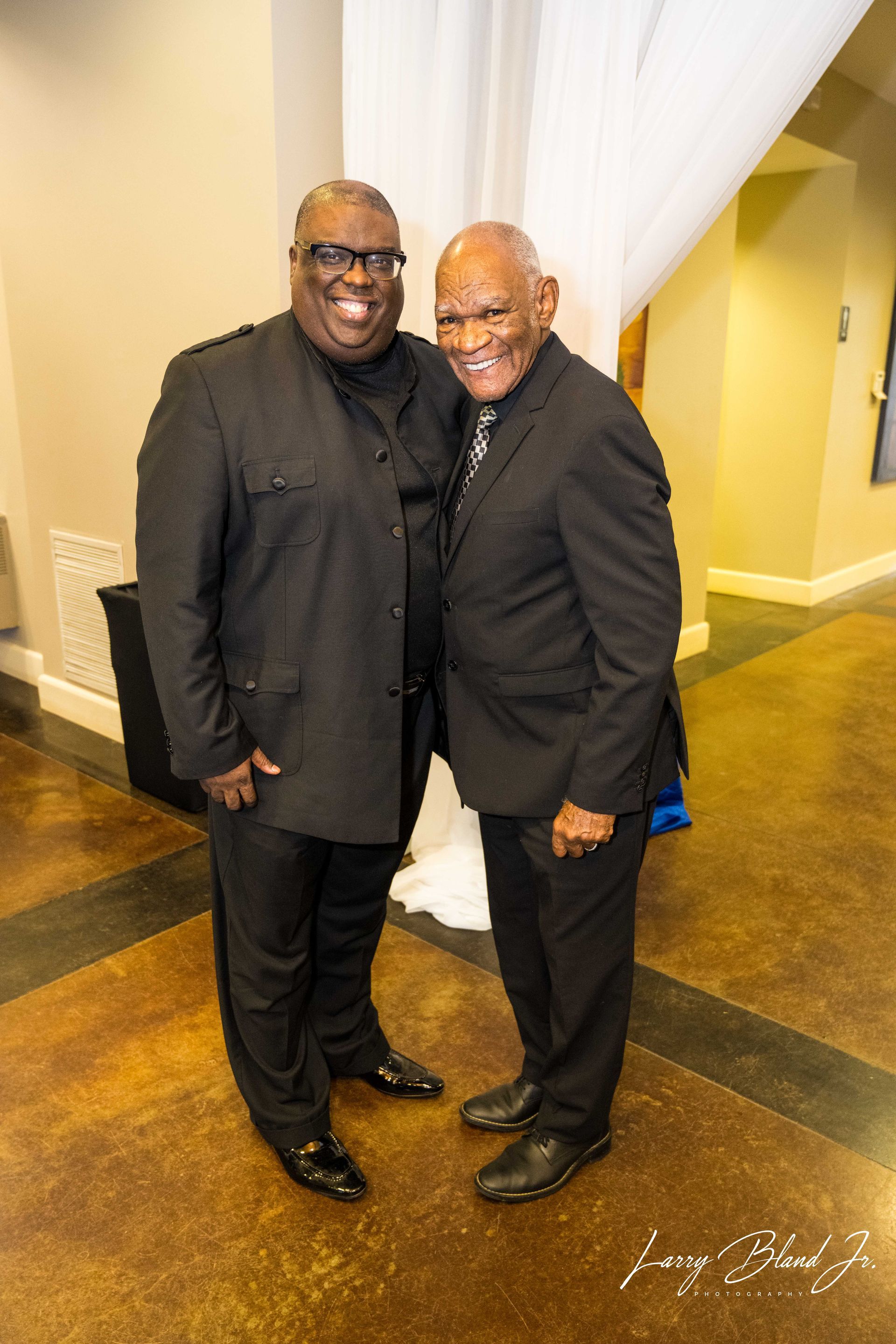 Two men in black suits smiling, posing together indoors near a white curtain.