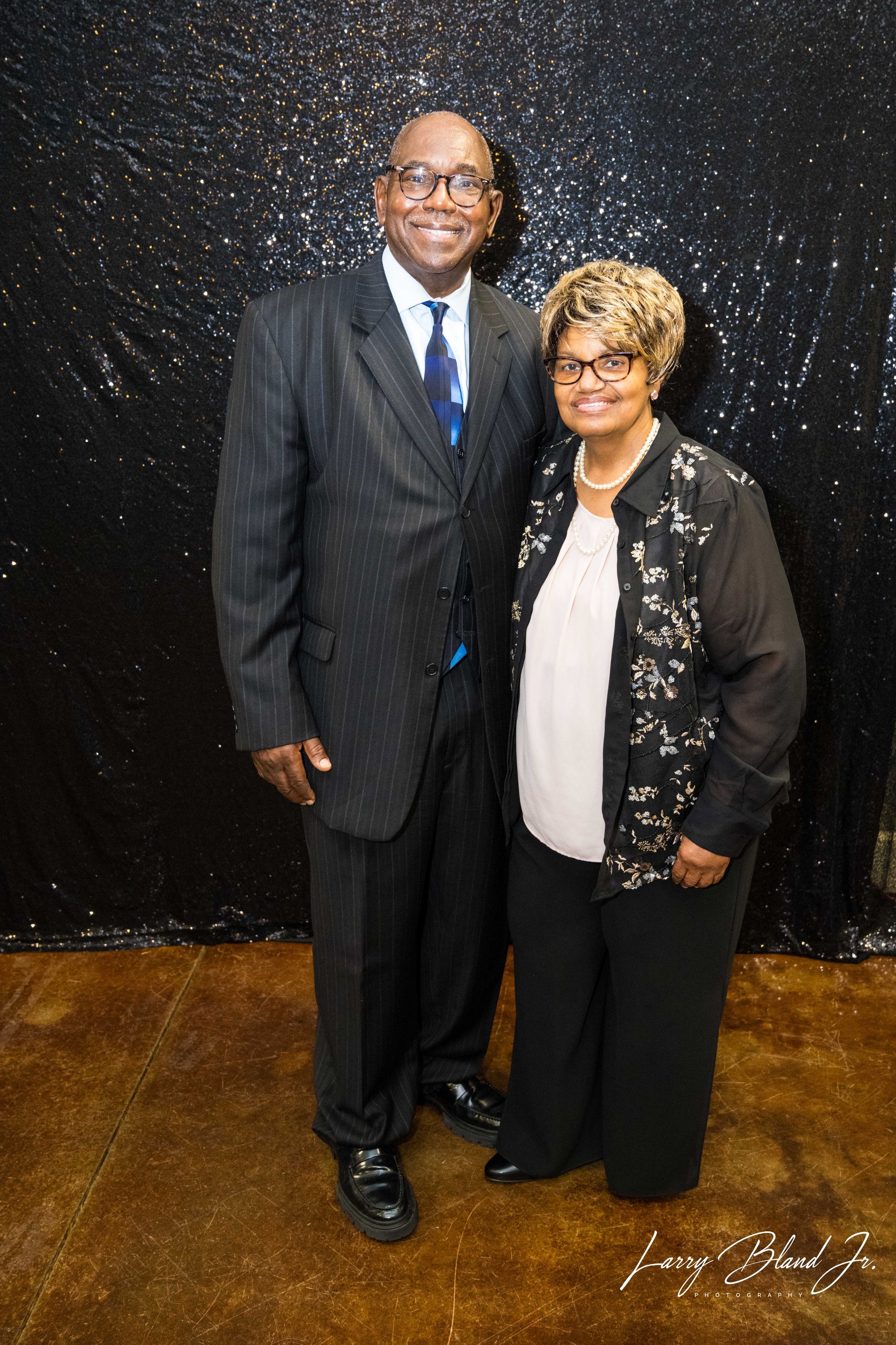 Man in suit and woman in black outfit smiling, posing together against a black sequin backdrop.