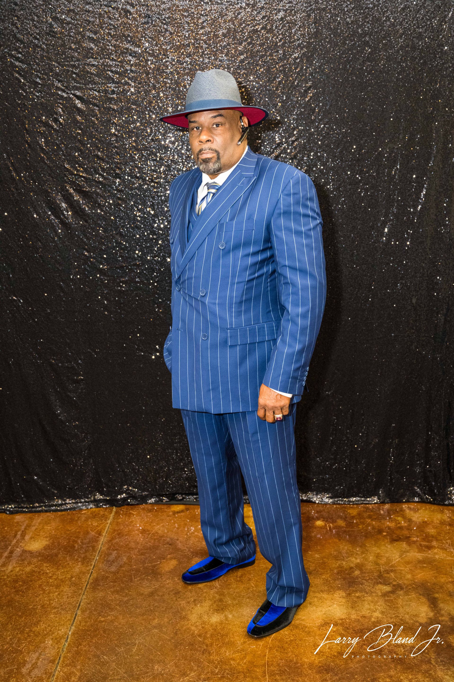 Man in a blue pinstripe suit and fedora stands in front of a sparkly black backdrop.